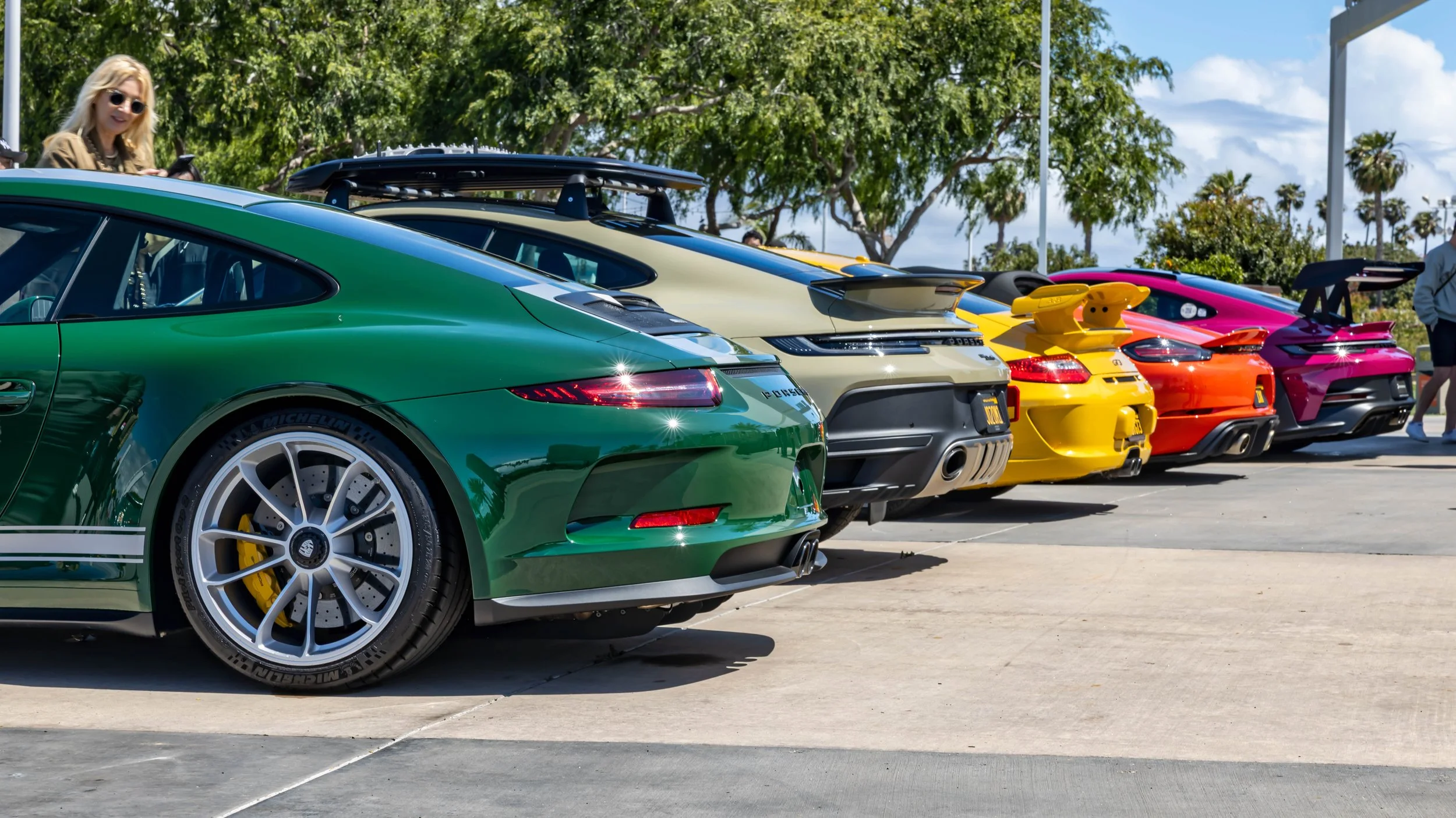 Air | Water 2025 — Orange County Fairgrounds — A low-angle, rear three-quarter view of a colorful lineup of modern Porsche sports cars parked in a row on a sunny day. The primary subject is a green Porsche 911 GT3 Touring with silver center-lock whee