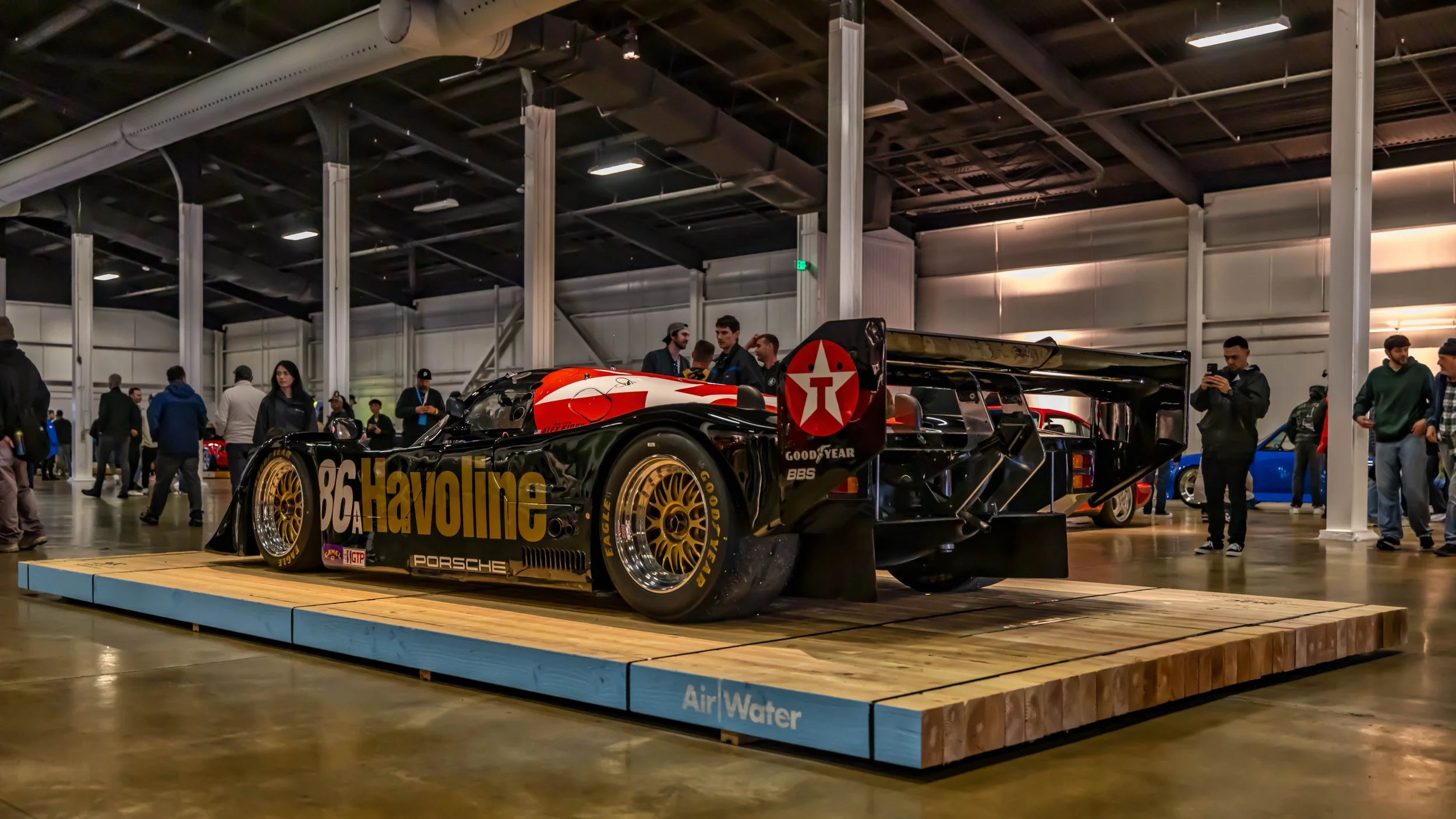 Air | Water 2025 — Orange County Fairgrounds — A low-angle, rear three-quarter view of a number 86 Porsche 962 race car in a black, red, and white Havoline/Texaco livery. The car is displayed on a wooden platform at the "Air | Water" indoor car show,