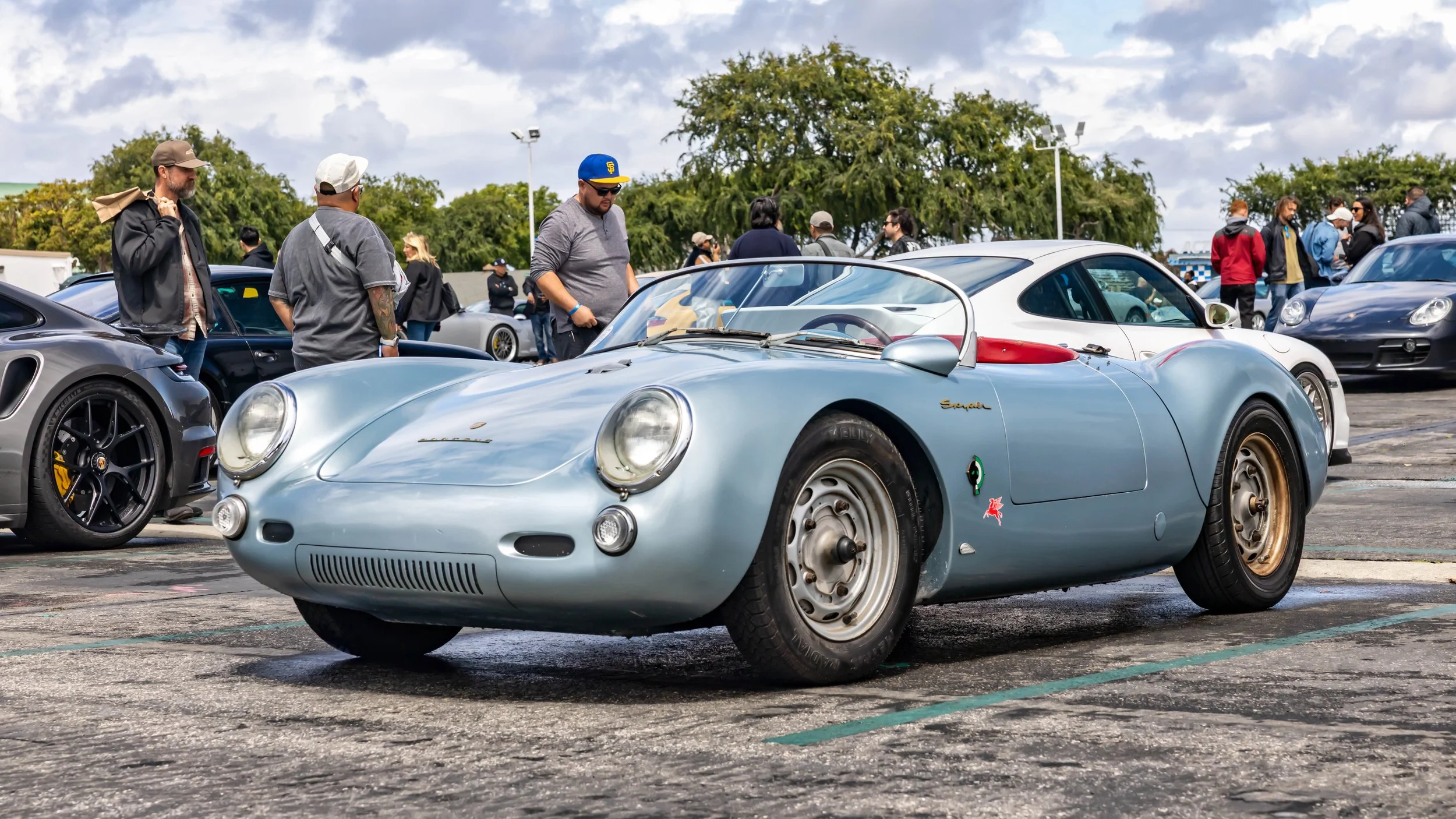 Air | Water 2025 — Orange County Fairgrounds — A low-angle, front three-quarter view of a vintage light blue Porsche 550 Spyder parked on asphalt at an outdoor car gathering. The car features simple steel wheels, a low windscreen, and a red Pegasus d