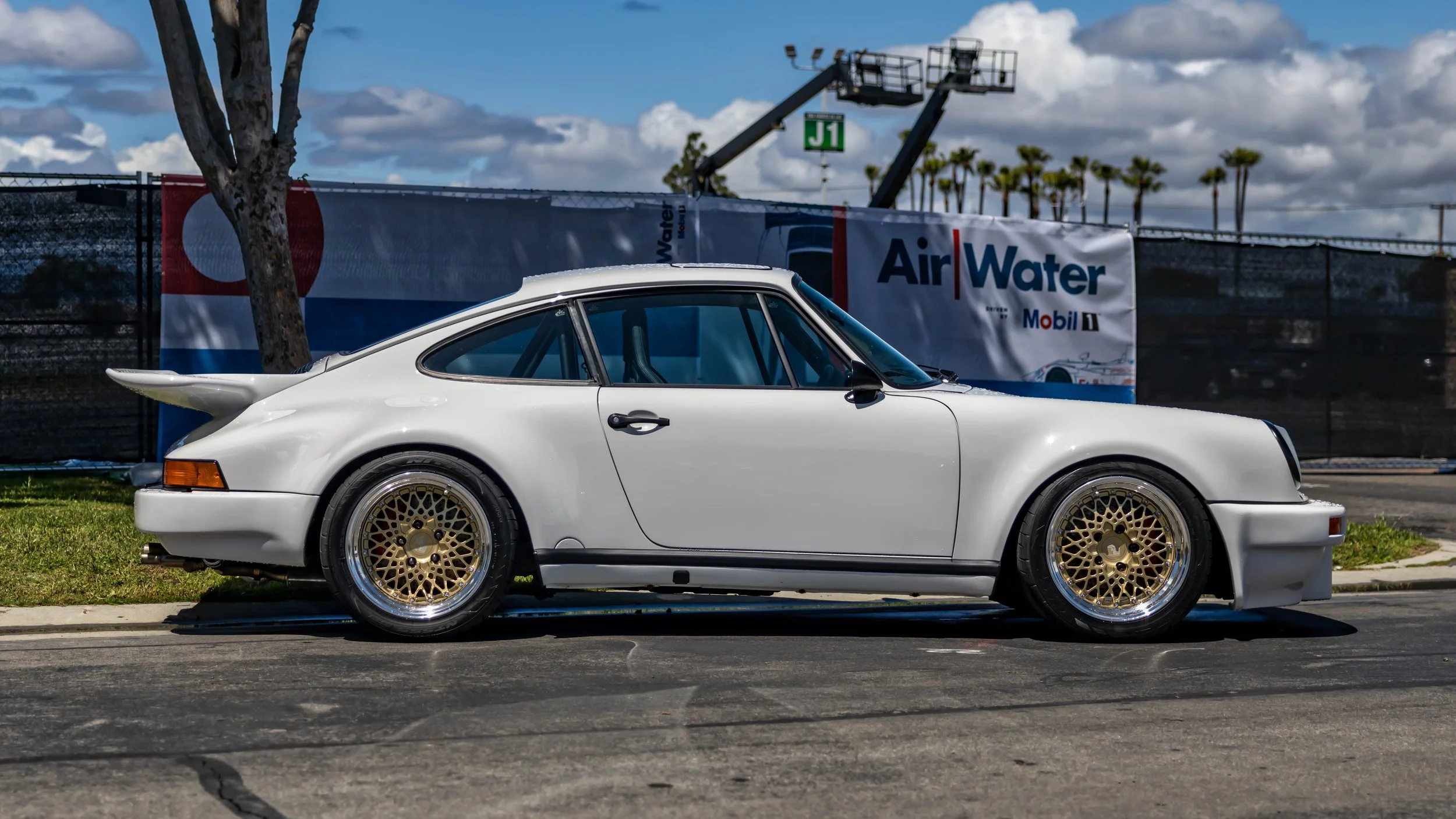 Air | Water 2025 — Orange County Fairgrounds — A low-angle, side profile photograph of a white 1980s Porsche 911 Turbo parked on an asphalt lot. The car is modified with gold BBS-style mesh wheels and has a roll cage visible through the window. In th