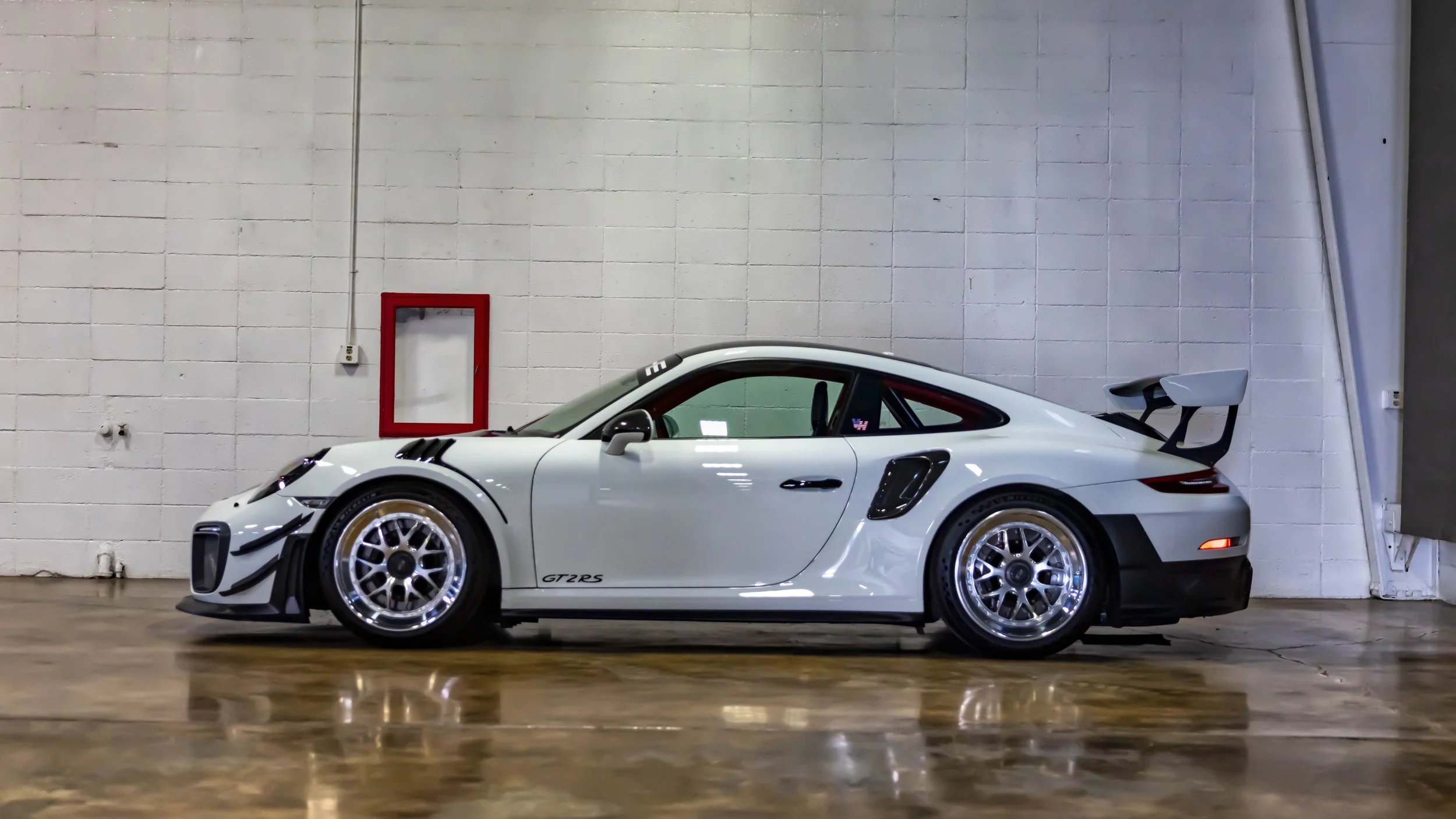 Air | Water 2025 — Orange County Fairgrounds — A white Porsche 911 GT2 RS (991.2) is captured in a side profile view, parked inside a garage or workshop. The vehicle is modified with aftermarket multi-piece wheels featuring polished lips, a visible r
