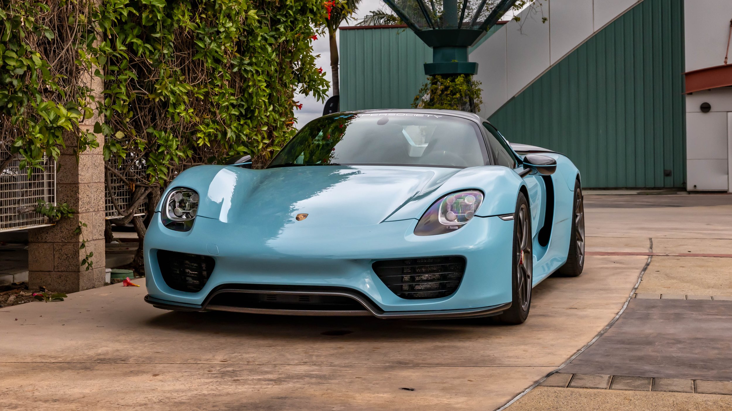 Air | Water 2025 — Orange County Fairgrounds — A light blue Porsche 918 Spyder is parked at a front three-quarter angle on a concrete driveway. The hypercar features red brake calipers and a 'SUPERSOCIETY' windshield banner. To the left is a stone pi