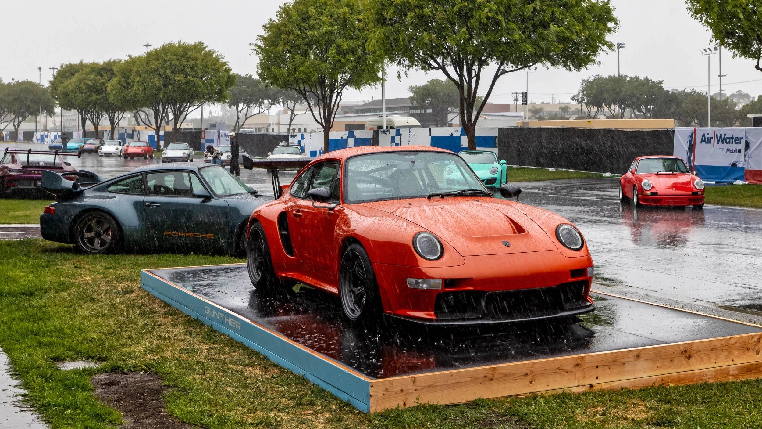 Air | Water 2025 — Gunther Werks — Orange County Fairgrounds — An orange Gunther Werks Turbo (Tornado) remastered Porsche 993 Remastered is showcased on a wet, wood-framed display platform during a heavy downpour. To its left, a dark green Porsche 99