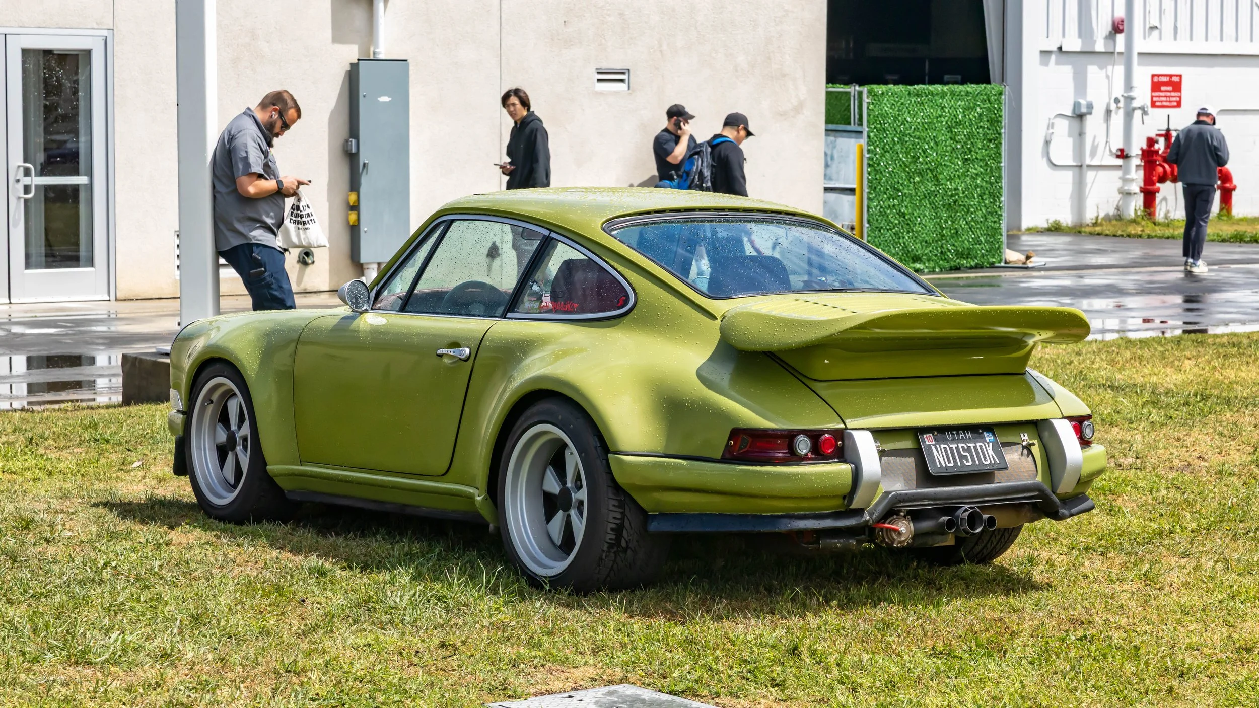 Air | Water 2025 — Orange County Fairgrounds — A rear three-quarter view of a heavily modified, lime green classic Porsche 911 parked on a wet lawn. The car features wide fender flares, a large 'whale tail' spoiler, silver and black Fuchs-style wheel