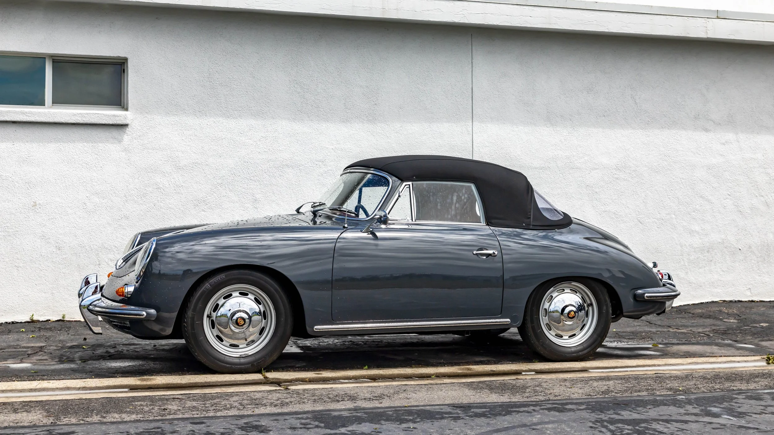 Air | Water 2025 — Orange County Fairgrounds — A vintage Porsche 356C Cabriolet, finished in a dark Slate Grey with its black soft top raised, is parked in profile against a white stucco wall. The car features chrome bumpers with over-riders and stee