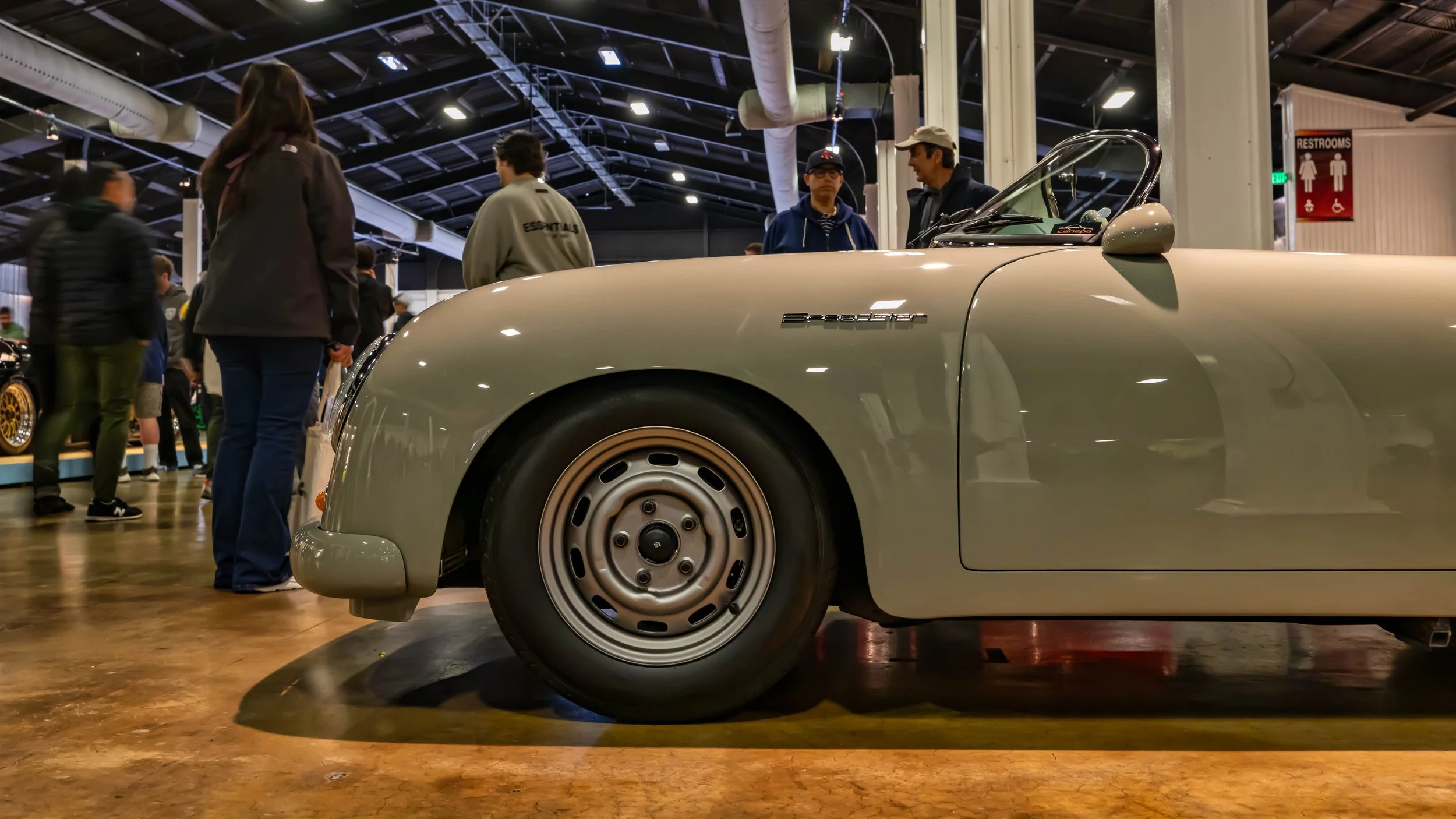 Air | Water 2025 — Orange County Fairgrounds — A low-angle, passenger-side profile view of a classic Porsche 356 Speedster finished in a light grey color. The car is displayed indoors on a polished concrete floor at a car show, with attendees visible