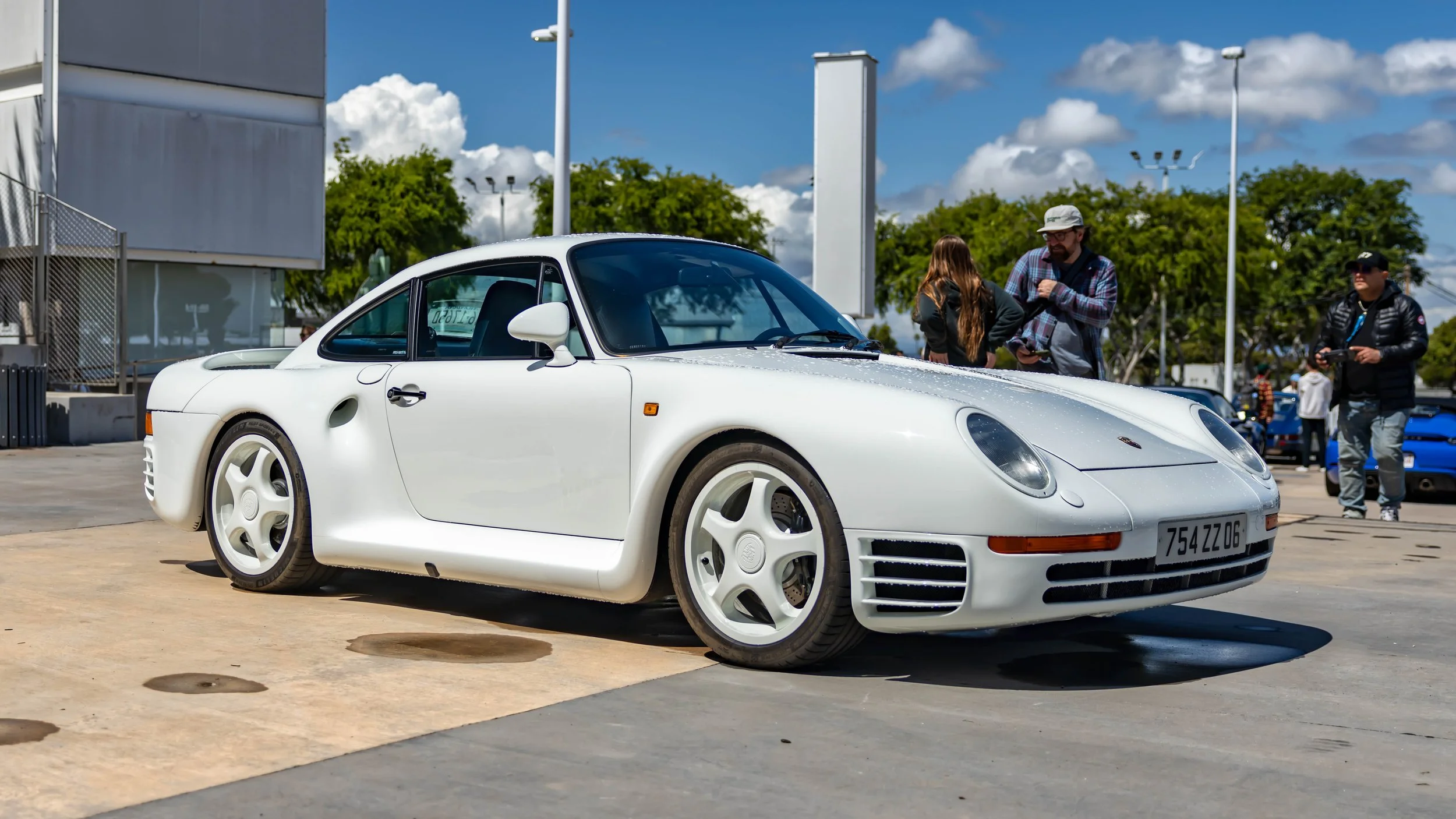 Air | Water 2025 — Orange County Fairgrounds — A white Porsche 959 is parked on a concrete lot under a bright, sunny sky. The car is shown from a low, front three-quarter angle, highlighting its iconic hollow-spoke magnesium wheels and integrated rea
