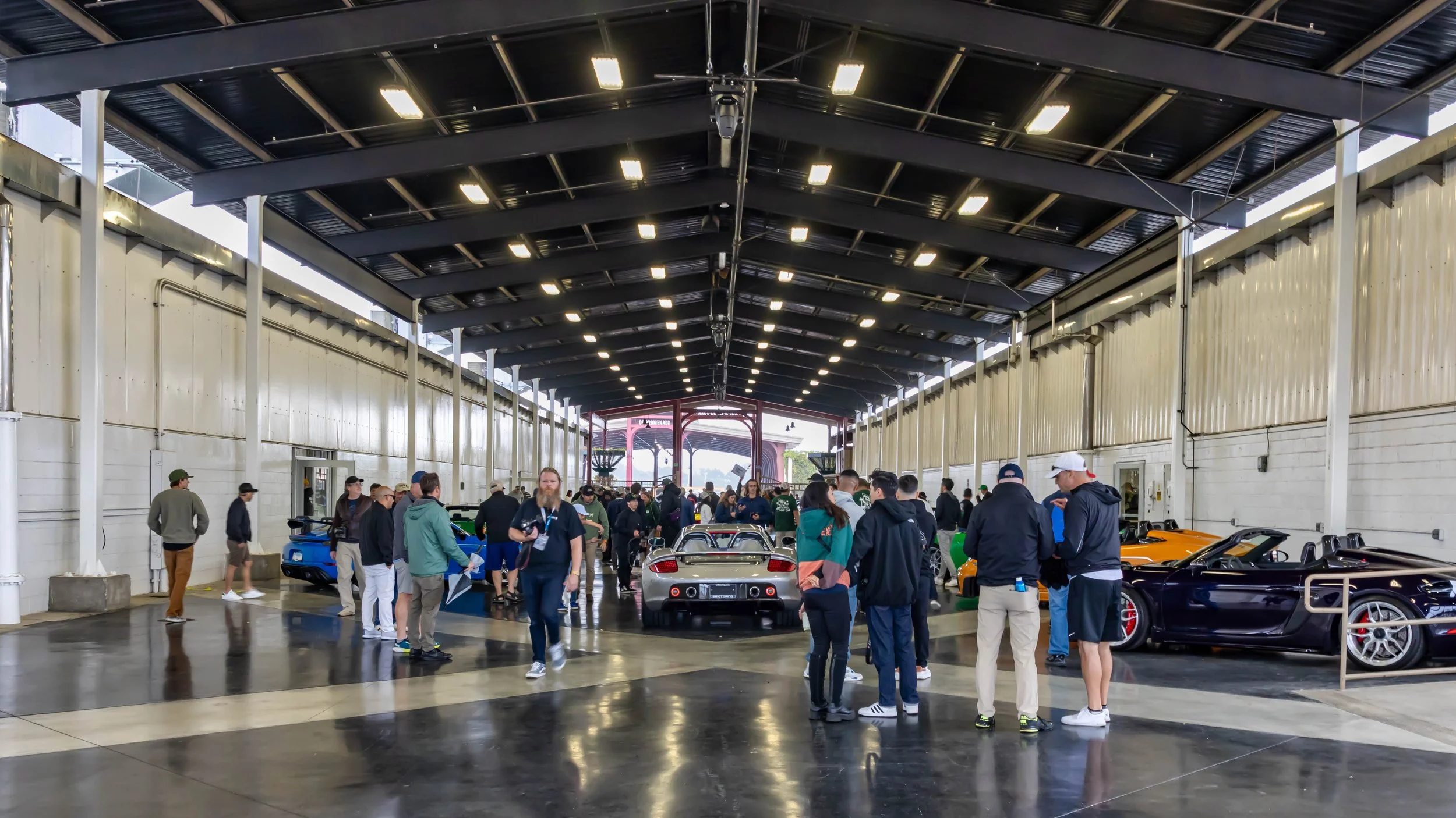 Air | Water 2025 — Orange County Fairgrounds — A wide, eye-level view of an large, open-sided exhibition hall. A silver Porsche Carrera GT is the central focus, parked on a reflective polished concrete floor. Numerous attendees are gathered around, a