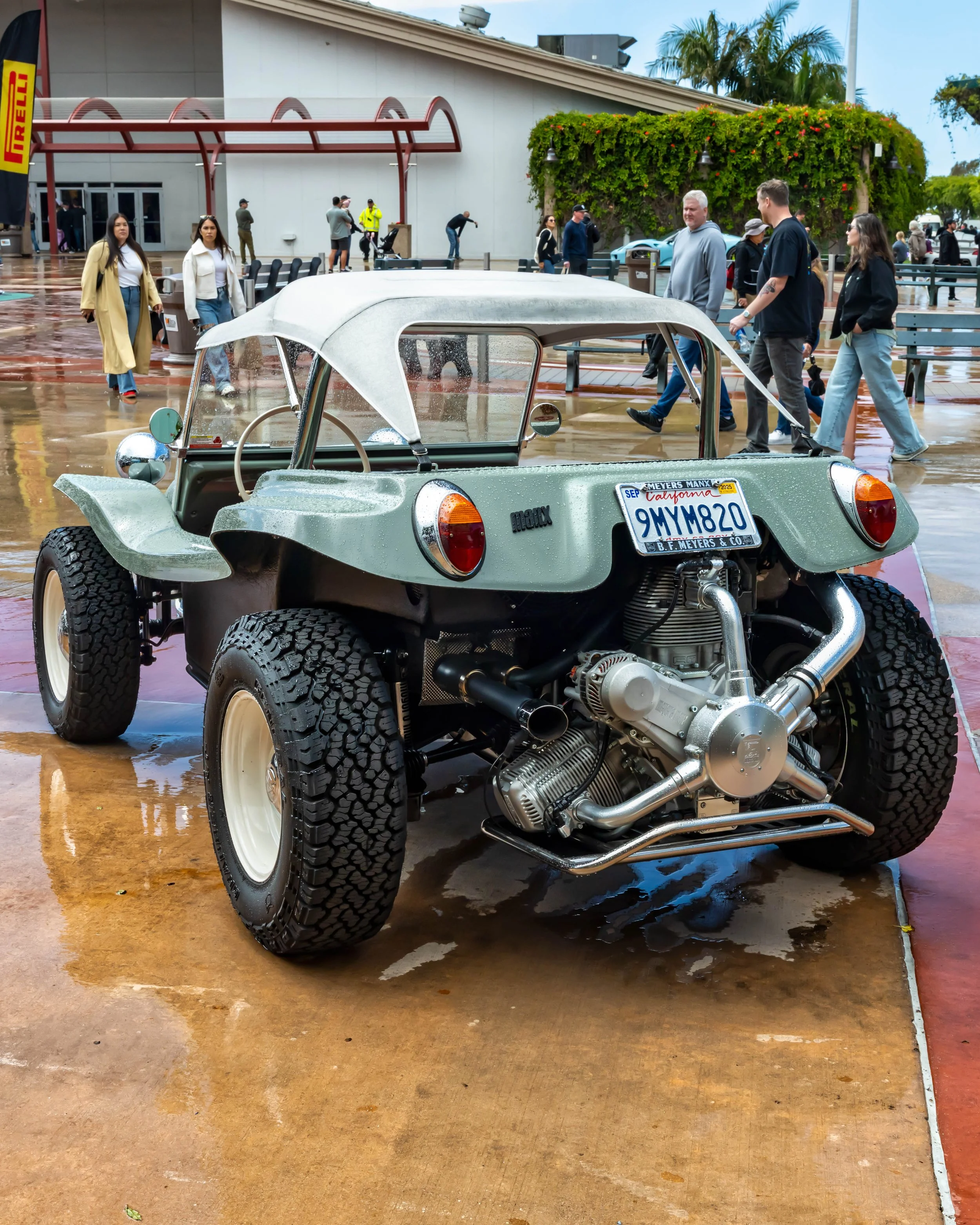 Air | Water 2025 — Orange County Fairgrounds — A rear three-quarter view of a vintage sage green Meyers Manx dune buggy parked on wet pavement at an outdoor event. The buggy features a white soft top, exposed rear engine, and chunky all-terrain tires