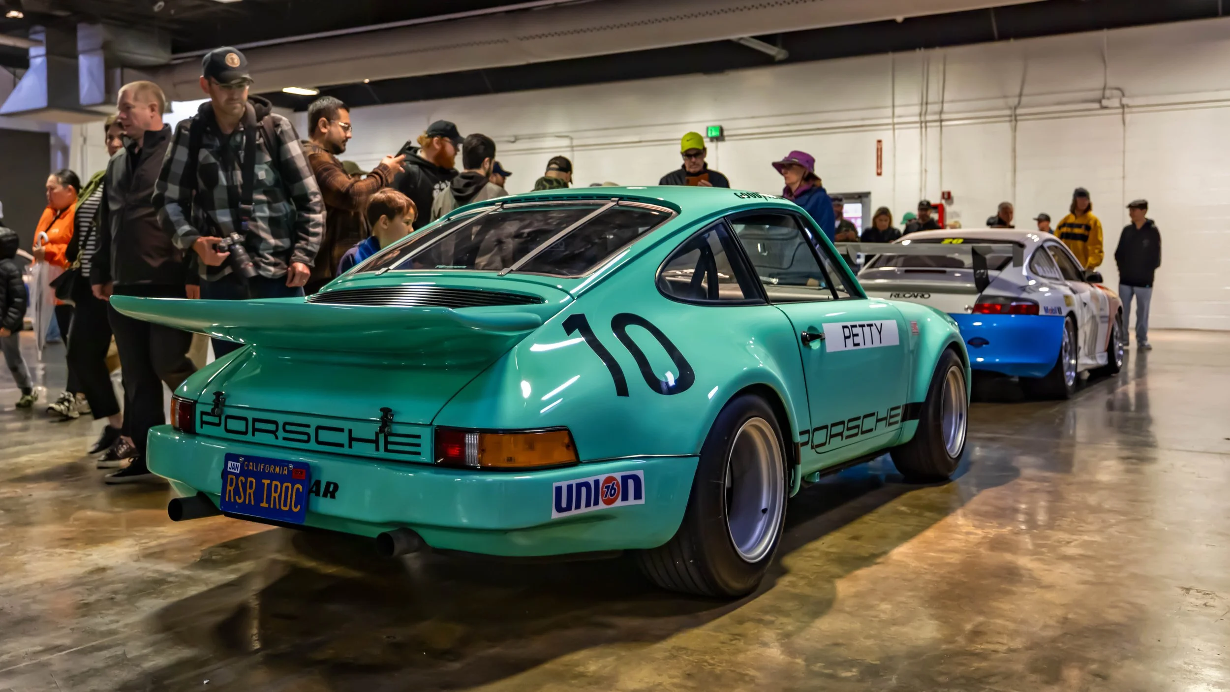 Air | Water 2025 — Orange County Fairgrounds — A low-angle, rear three-quarter view of a seafoam green Porsche 911 RSR IROC tribute car, featuring the number "10" and "PETTY" livery. The car is displayed indoors on a polished concrete floor at a car 