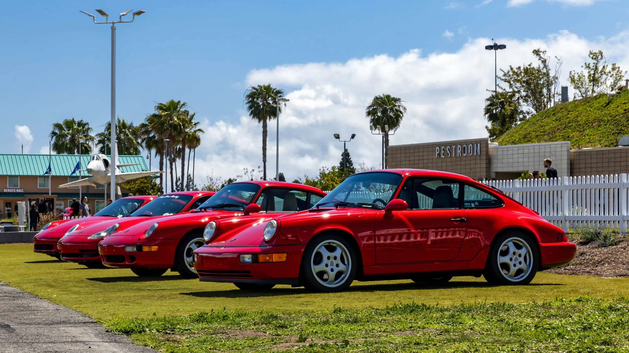 Air | Water 2025 — Orange County Fairgrounds — A lineup of four red Porsche 911s is parked on a manicured grass lawn under a partly cloudy sky. The primary subject, a 964-generation Carrera in the foreground, is shown from a front three-quarter view,