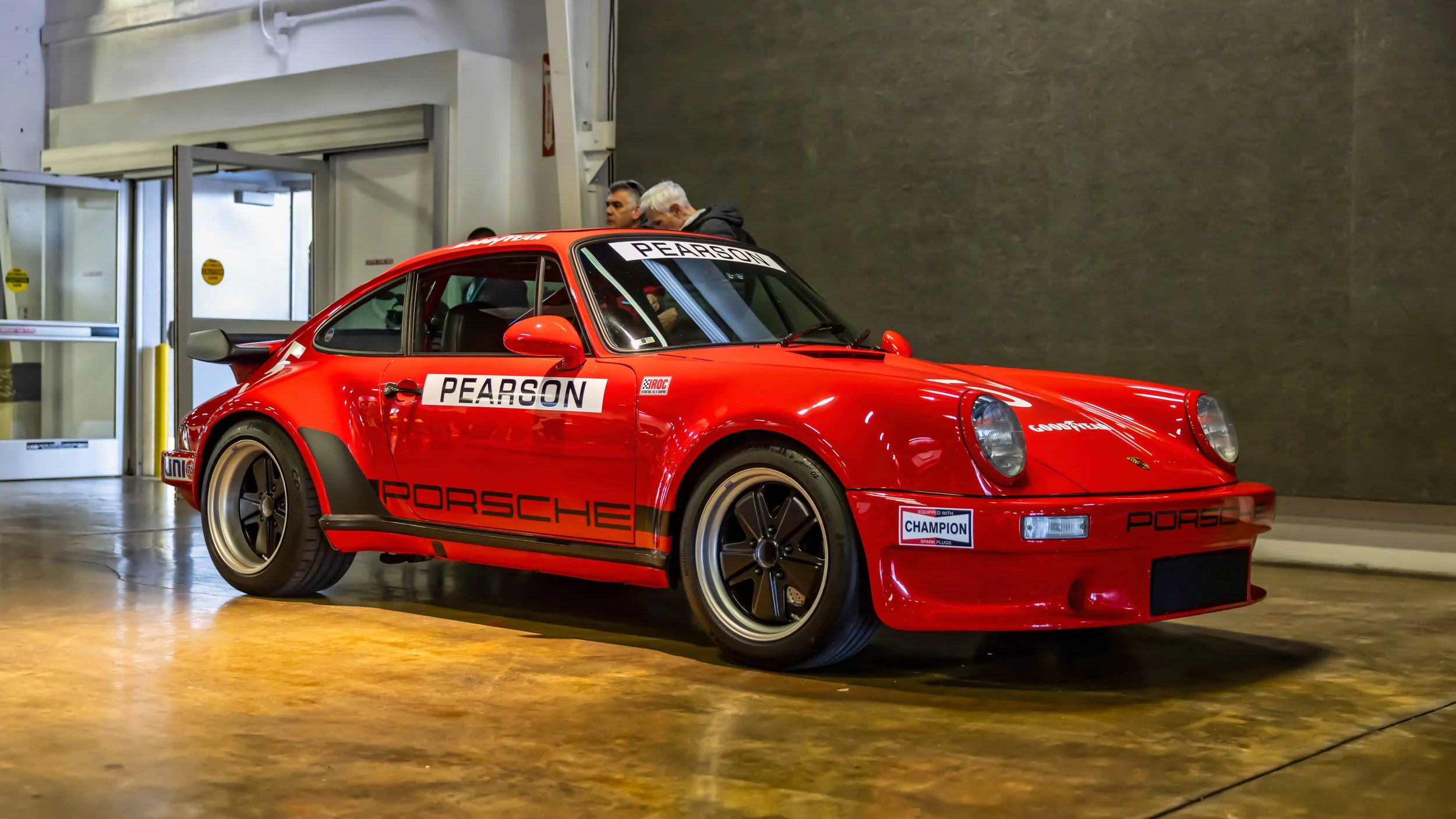 Air | Water 2025 — Orange County Fairgrounds — A bright red 1974 Porsche 911 Carrera RSR IROC tribute car is displayed indoors on a polished, reflective concrete floor. The car is adorned with a full racing livery, including 'PEARSON' on the doors an