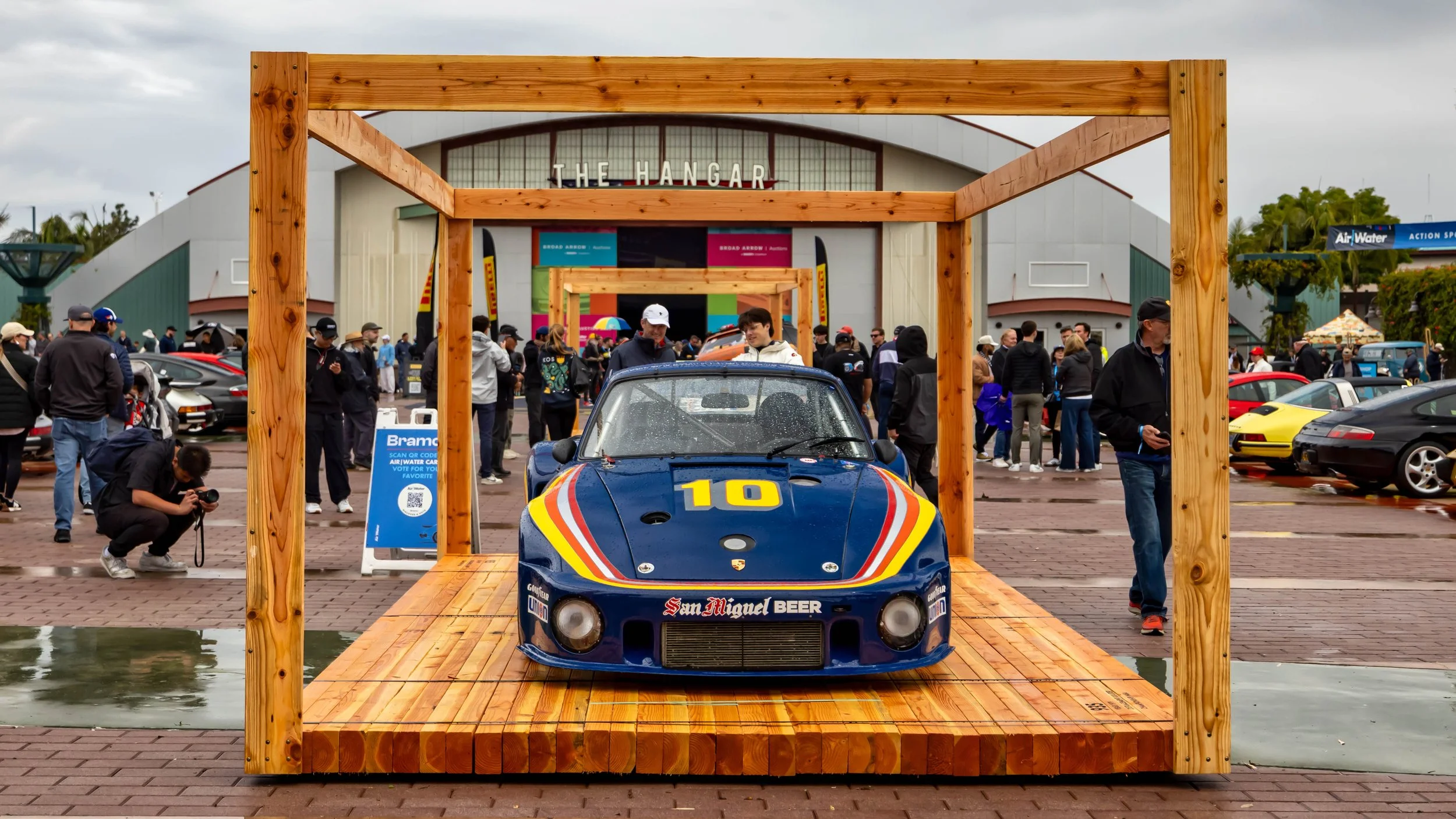 Air | Water 2025 — Orange County Fairgrounds — A front view of a vintage blue Porsche 935 race car, number 10, with a San Miguel Beer livery, displayed on a raised wooden platform. The event takes place on a wet, brick-paved lot in front of a large b