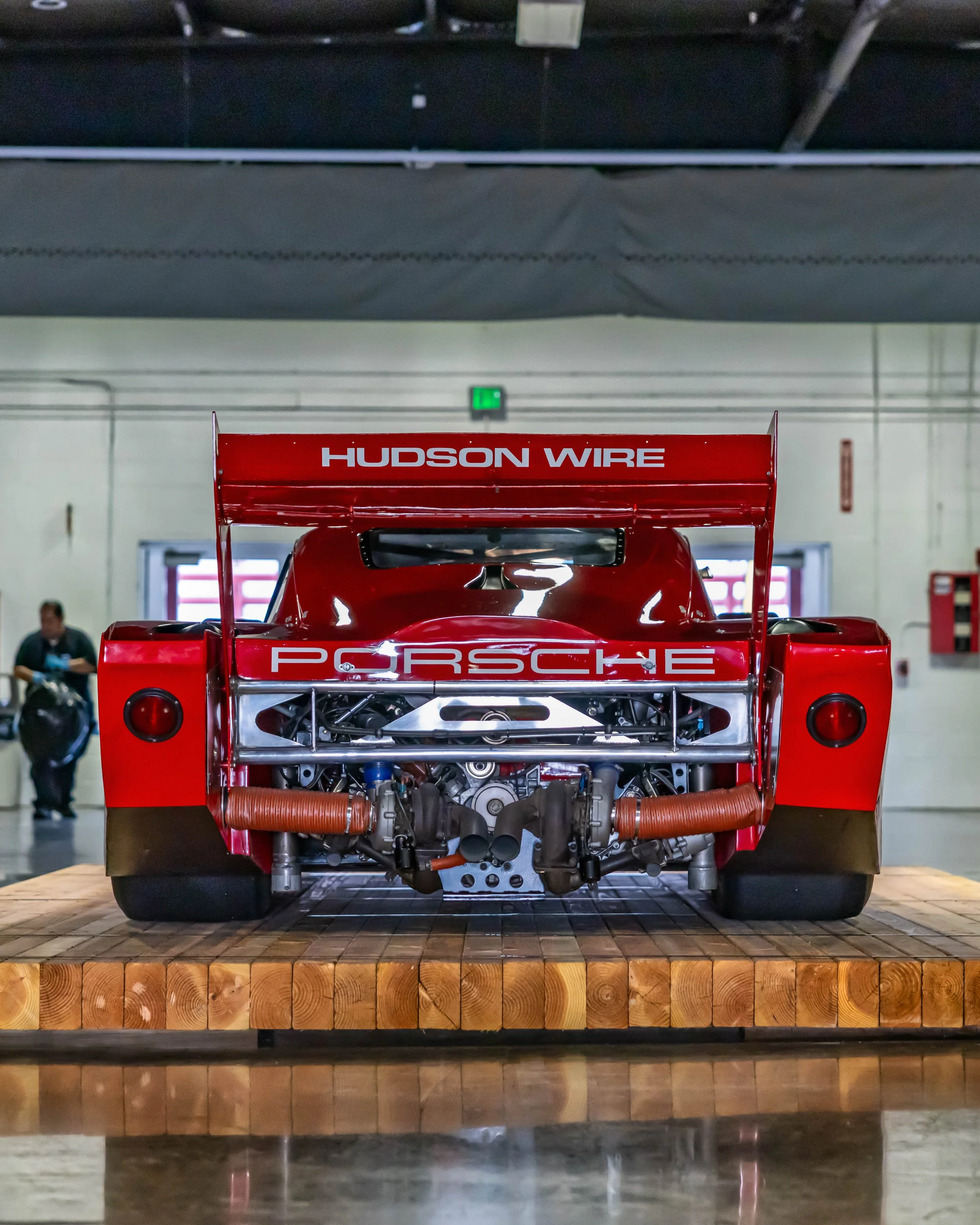 Air | Water 2025 — Orange County Fairgrounds — A rear view of a vintage red Porsche 935 K3 race car, presented on a heavy wooden block platform inside a large exhibition hall.. The car's wide bodywork, massive rear wing with 'HUDSON WIRE' livery, and