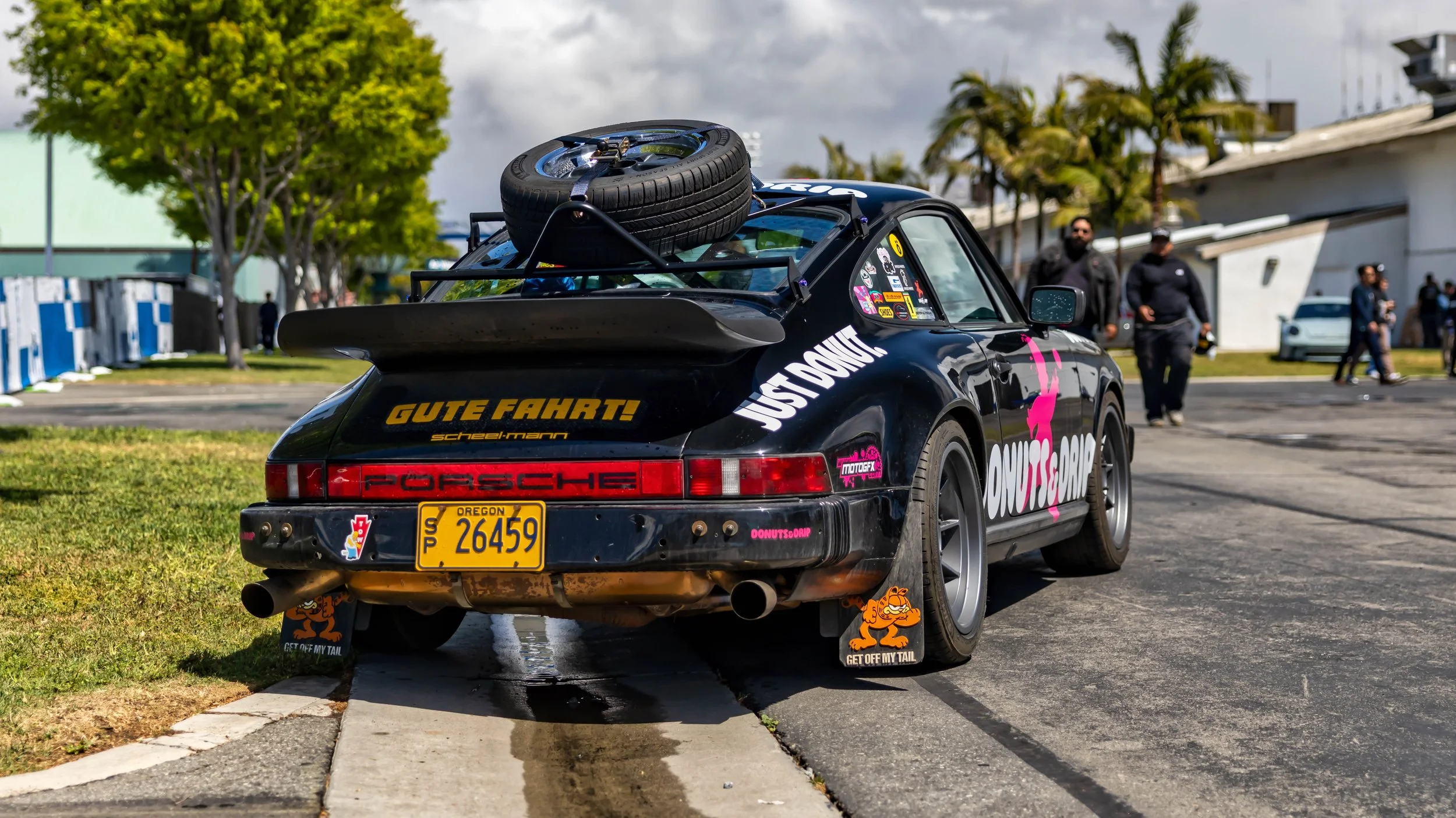 Air | Water 2025 — Orange County Fairgrounds — A modified black Porsche 911 G-body is parked on the edge of an asphalt lot, viewed from the rear three-quarter angle. The car features a large "whale tail" spoiler, a roof rack carrying a spare wheel an
