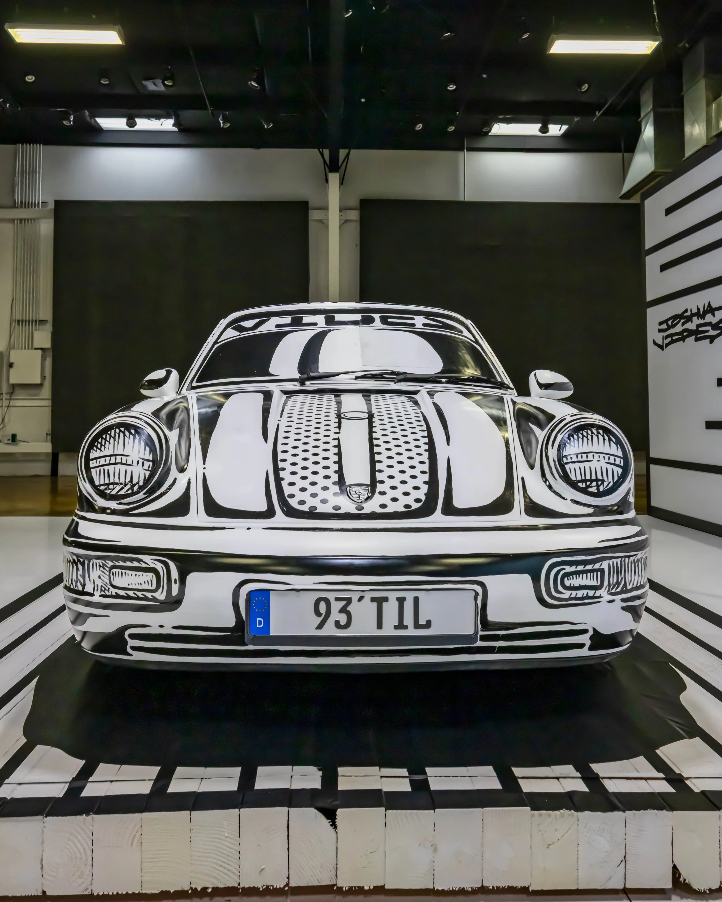 Air | Water 2025 — Orange County Fairgrounds — A front view of a Porsche 911 (993) art car by Joshua Vides, displayed indoors on a custom platform. The car is finished in a distinctive black-and-white, cartoon-like graphic style that outlines its fea