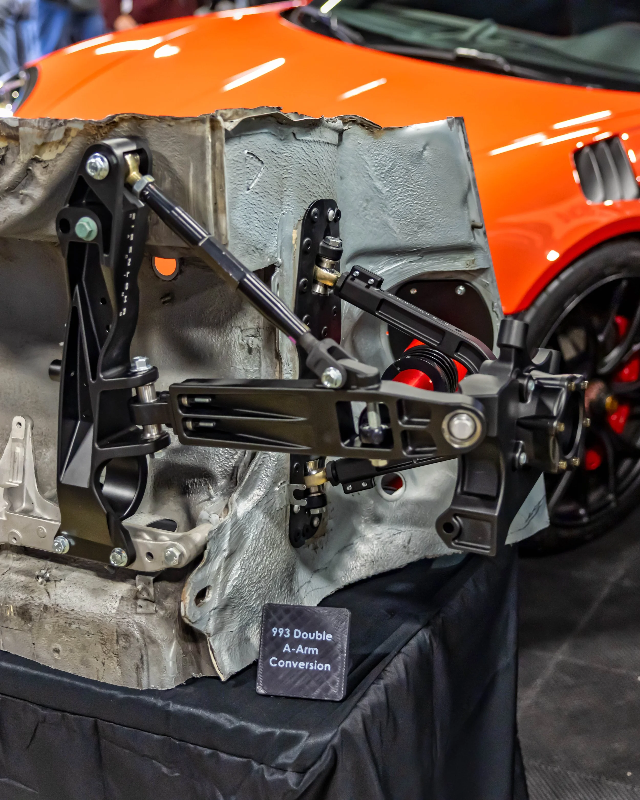 Air | Water 2025 — Orange County Fairgrounds — A detailed view of Eisenlohr Racing's double A-arm front suspension conversion for a Porsche 993, displayed on a cutaway chassis section at an indoor event. In the background, the front fender and wheel 
