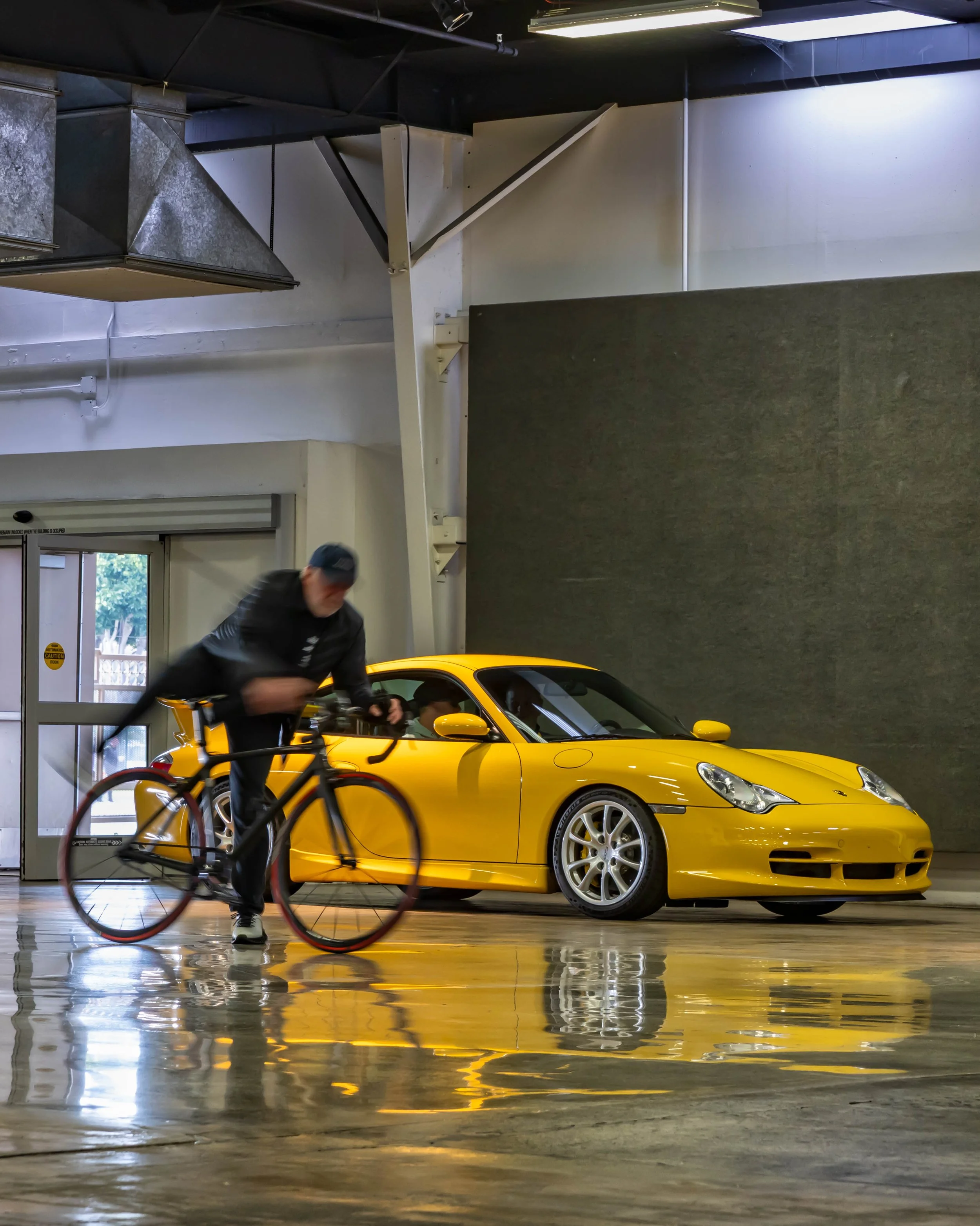 Air | Water 2025 — Orange County Fairgrounds — A Speed Yellow Porsche 996.2 911 GT3 is parked indoors on a highly reflective, polished concrete floor, creating a mirror-like effect. In the foreground, as Jeff Zwarts jumps on a bicycle is captured wit