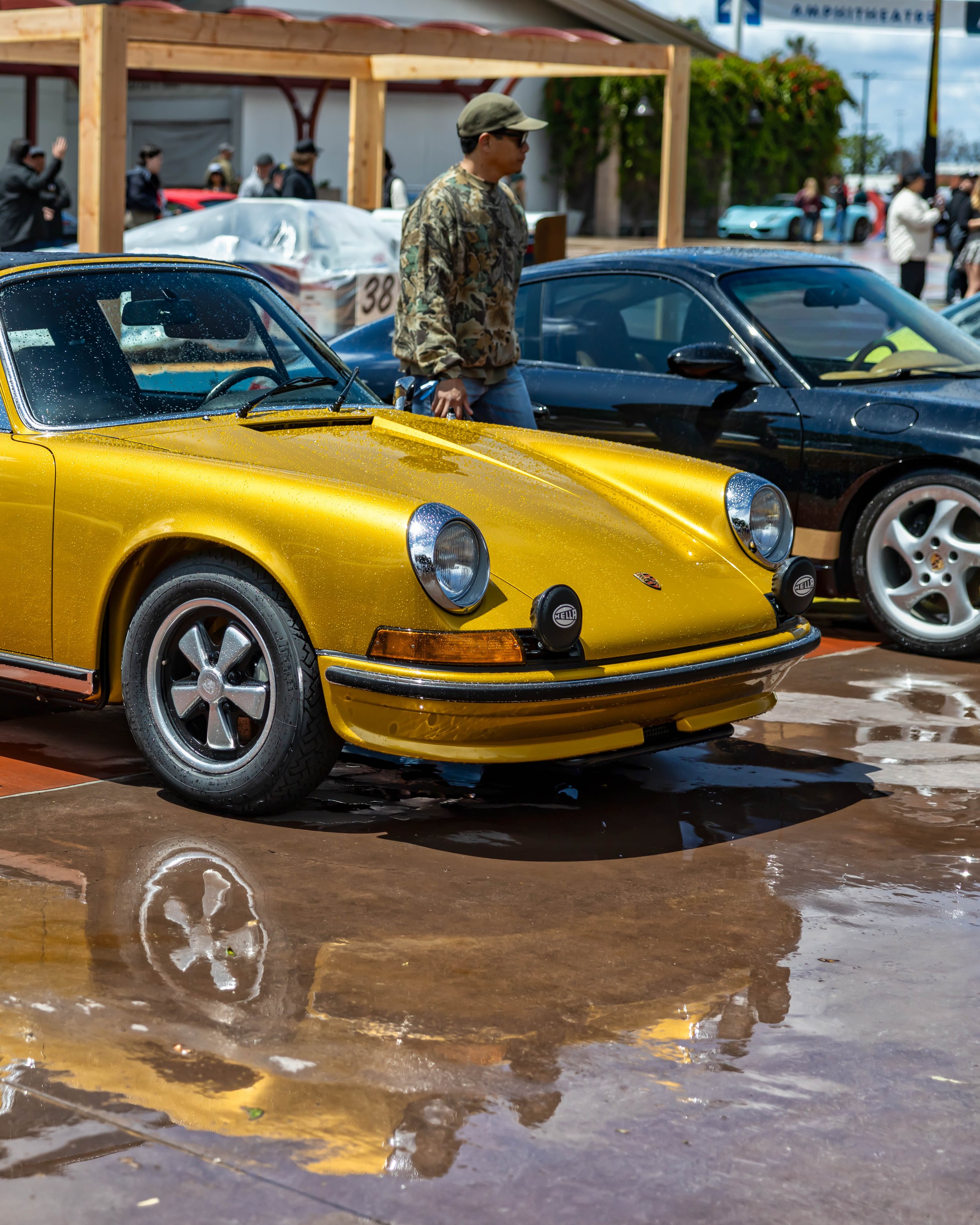 Air | Water 2025 — Orange County Fairgrounds — A front three-quarter view of a classic, long-hood Porsche 911 in a vibrant metallic yellow, covered in water droplets. The car, featuring Fuchs wheels and bumper-mounted Hella rally lights, is parked on