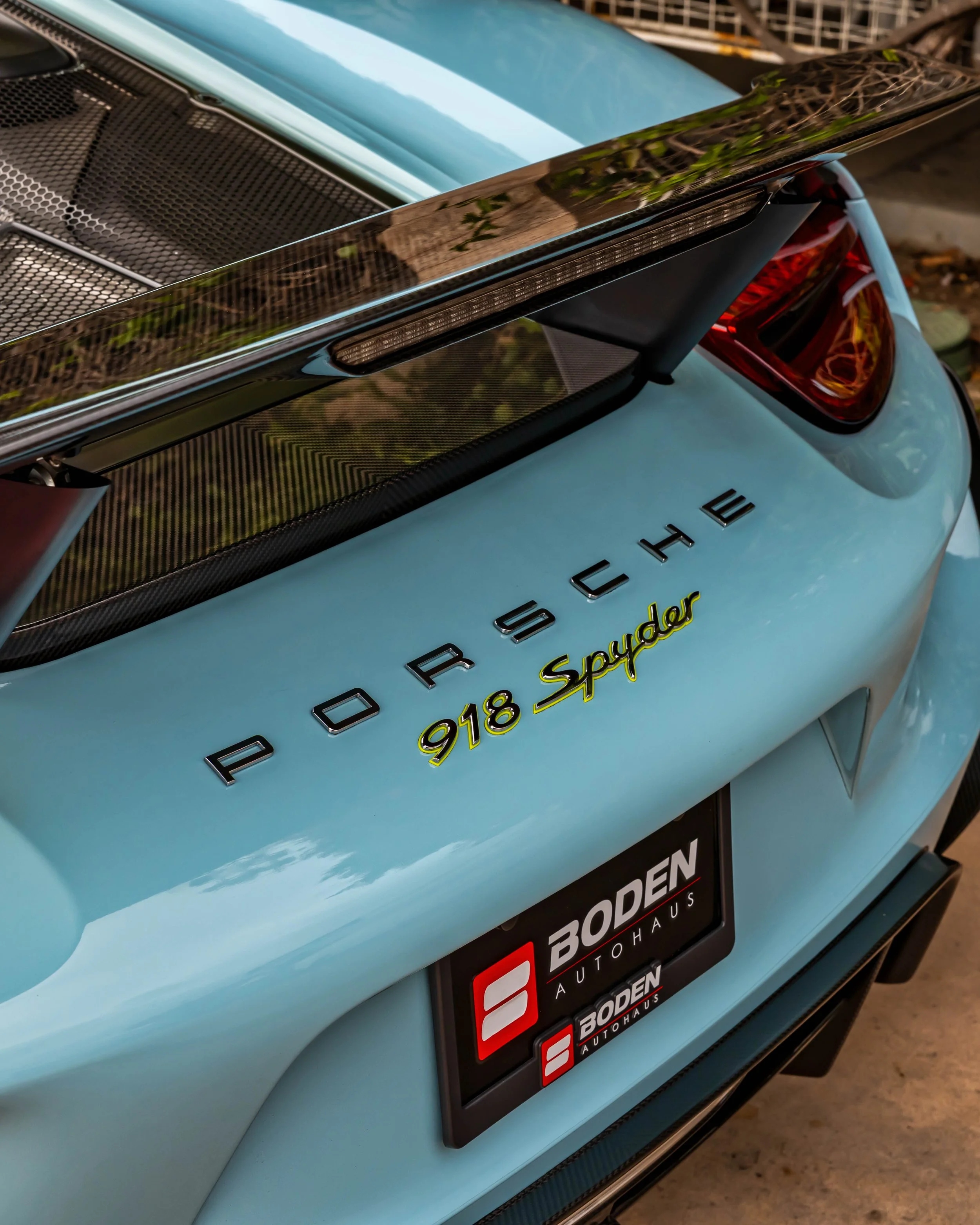 Air | Water 2025 — Orange County Fairgrounds — A high-angle, close-up detail shot of the rear of a light blue Porsche 918 Spyder. The image focuses on the 'PORSCHE' and '918 Spyder' badging on the decklid, with the latter featuring a yellow outline. 