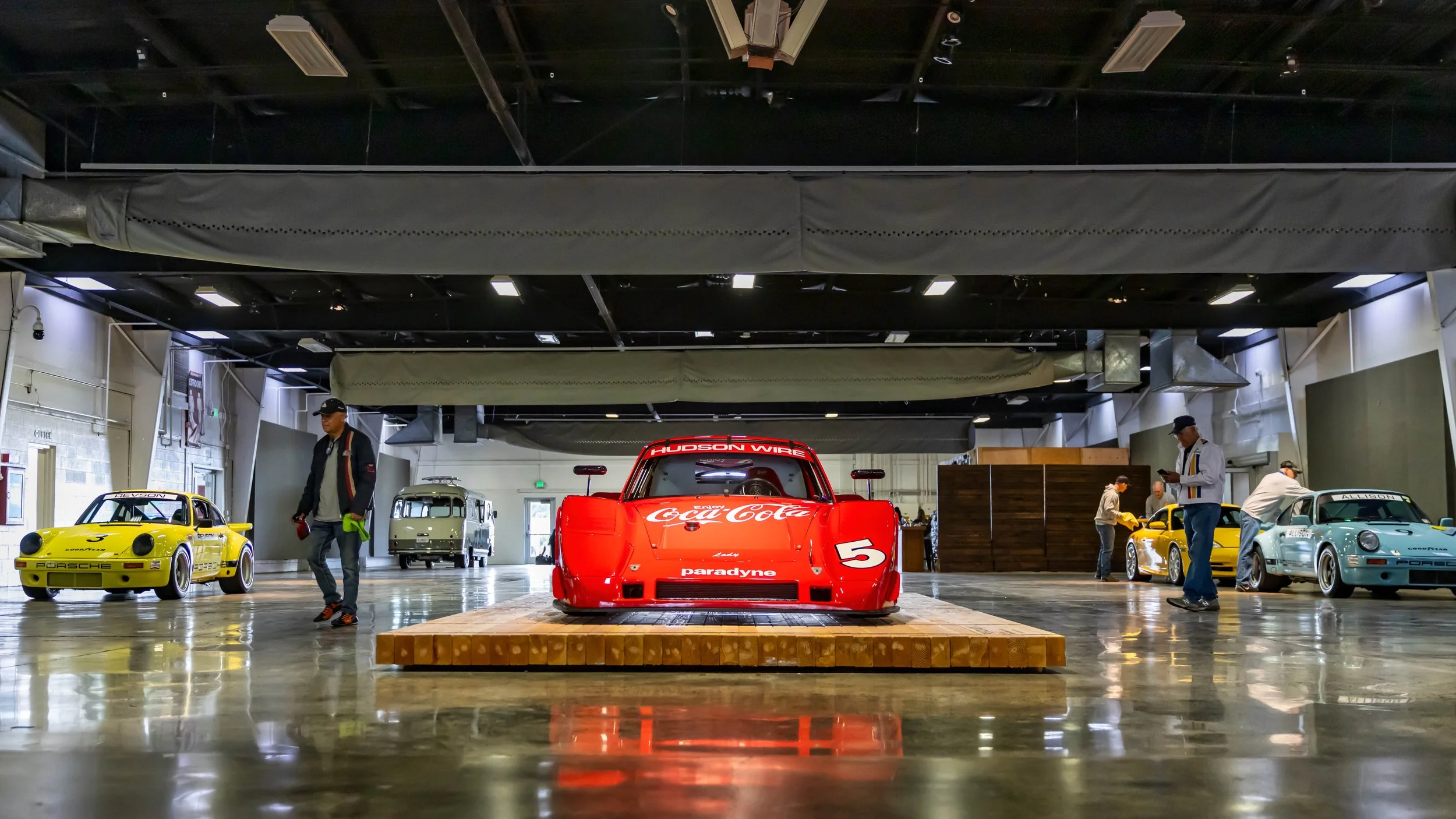 Air | Water 2025 — Orange County Fairgrounds — A low-angle, head-on photograph of a red #5 Porsche 935 K3 race car with Coca-Cola and Paradyne livery, centrally displayed on a wooden platform. The car is inside a large exhibition hall with a highly p