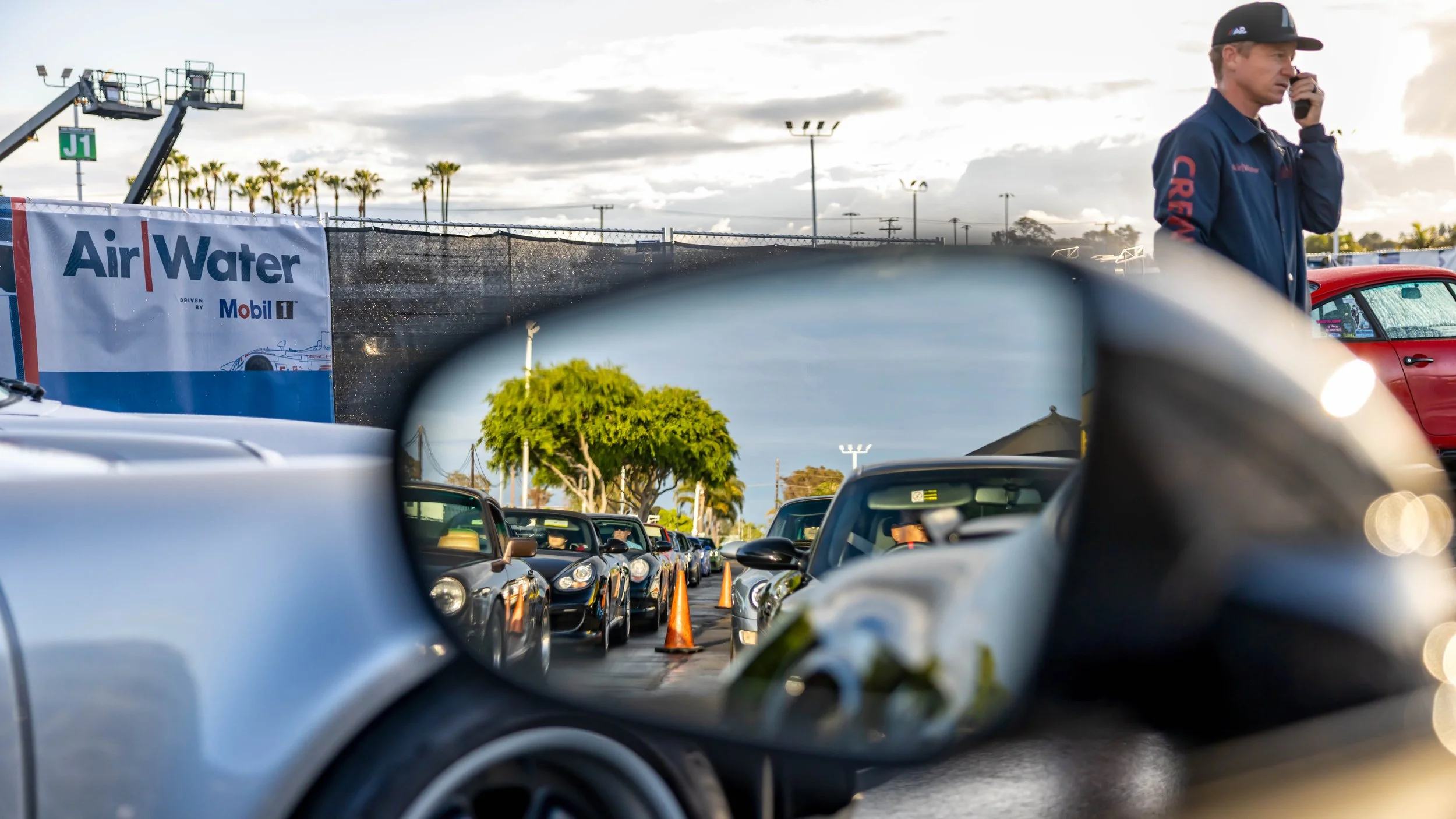 Air | Water 2025 — Orange County Fairgrounds — A creative shot from the driver's perspective of a silver Porsche, focusing on the side mirror which reflects a long queue of various Porsche models waiting to enter the 'Air | Water' car event. As Patri