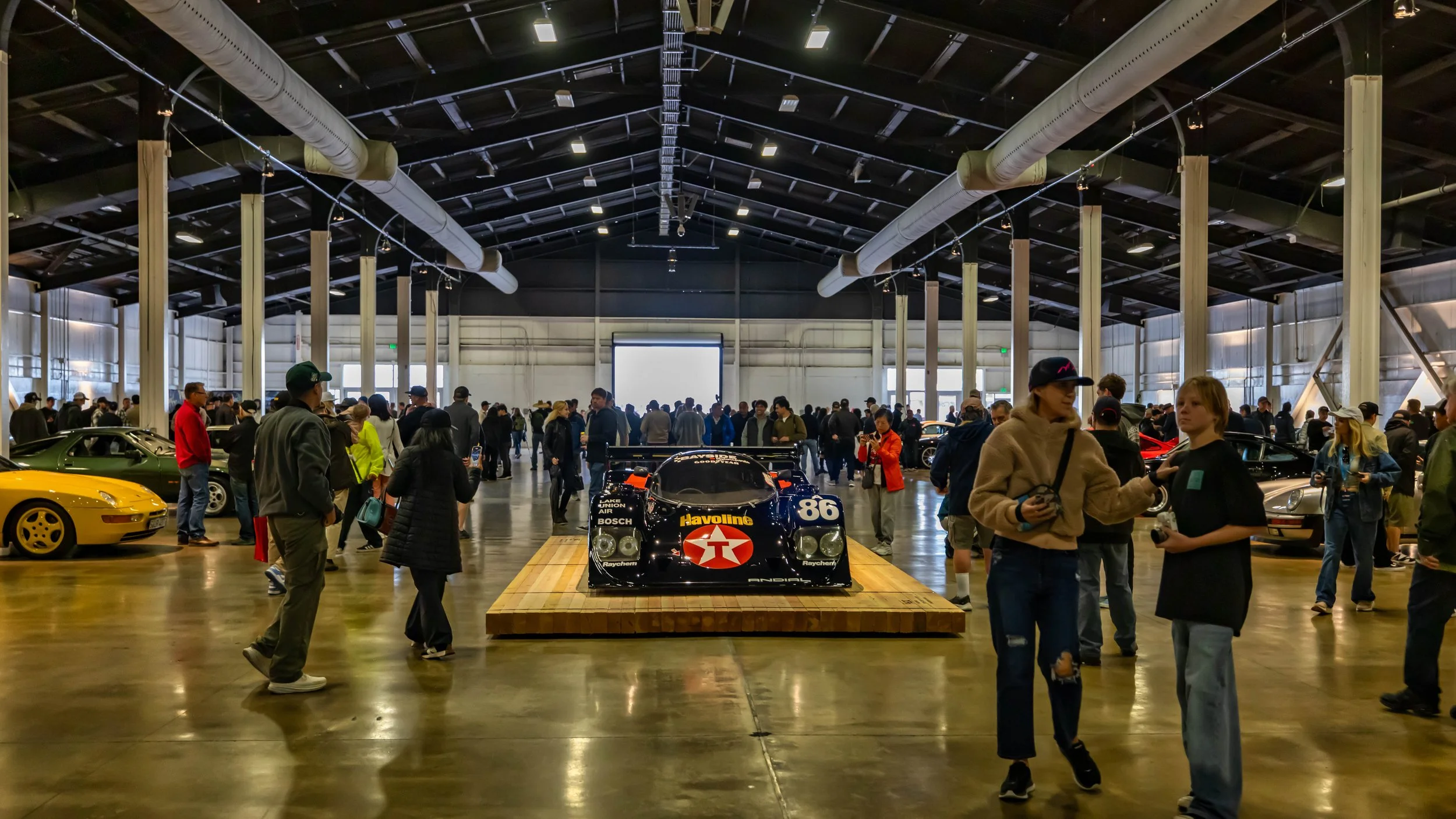 Air | Water 2025 — Orange County Fairgrounds — A wide, eye-level shot of a bustling indoor car show in a large exhibition hall, with a 1987 Porsche 962 race car as a centerpiece. The car, featuring a dark blue Havoline livery with the number 86, is e