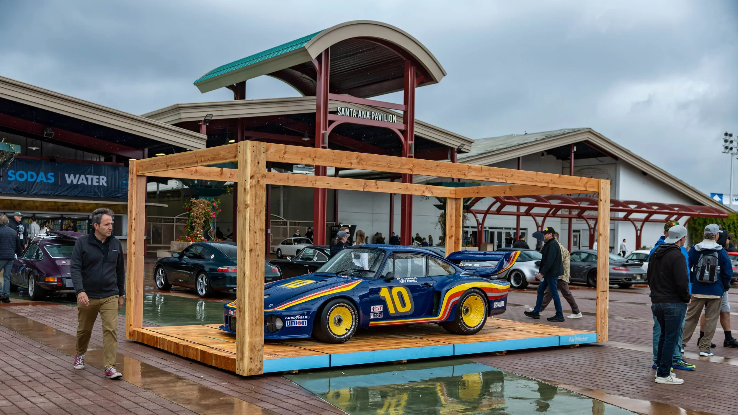 Air | Water 2025 — Orange County Fairgrounds — A blue 1979 Porsche 935 race car, number 10, with a distinctive red, orange, and yellow livery and yellow BBS turbo-fan wheels, is showcased on a wooden display platform. The scene is an outdoor car even