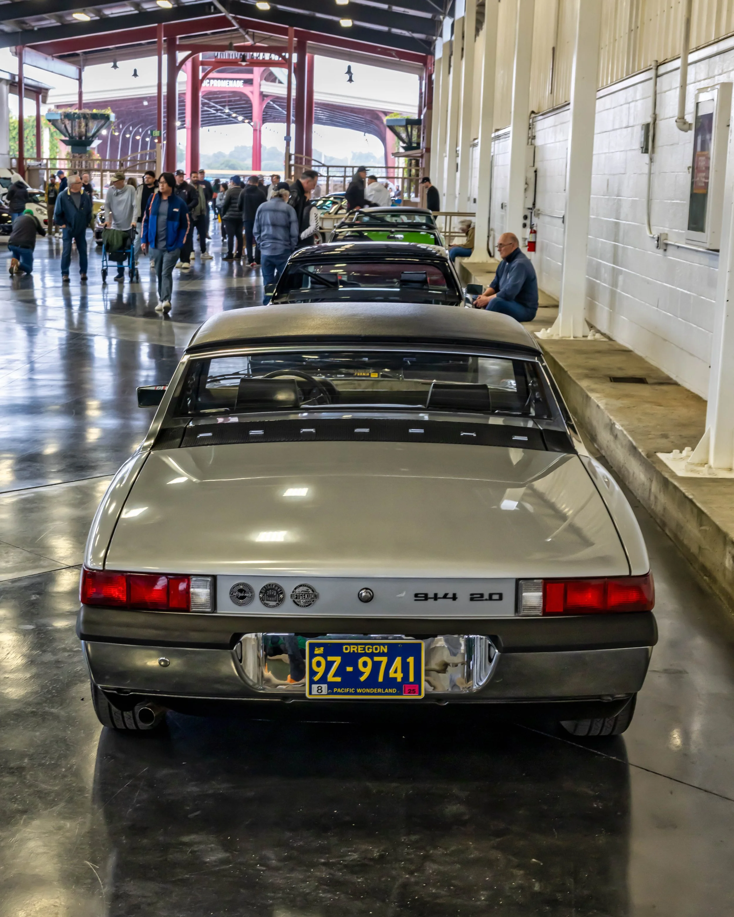Air | Water 2025 — Orange County Fairgrounds — A rear view of a silver Porsche 914 2.0 parked indoors on a reflective concrete floor during a car show. The venue is a large exhibition hall with red steel beams, and other classic cars and attendees ar