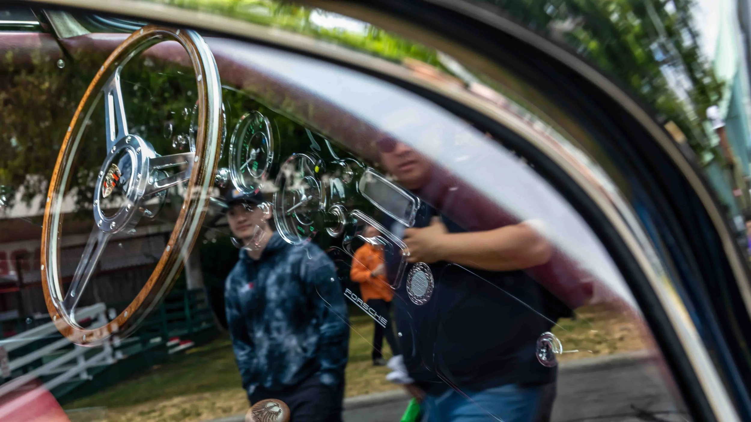 Air | Water 2025 — Emory Motorsports — OC Fairgrounds — A close-up view through the driver's side window of a classic Porsche 356 Emory Outlaw, focusing on the wood-rimmed steering wheel and green-faced gauges. The curved glass reflects the outdoor c