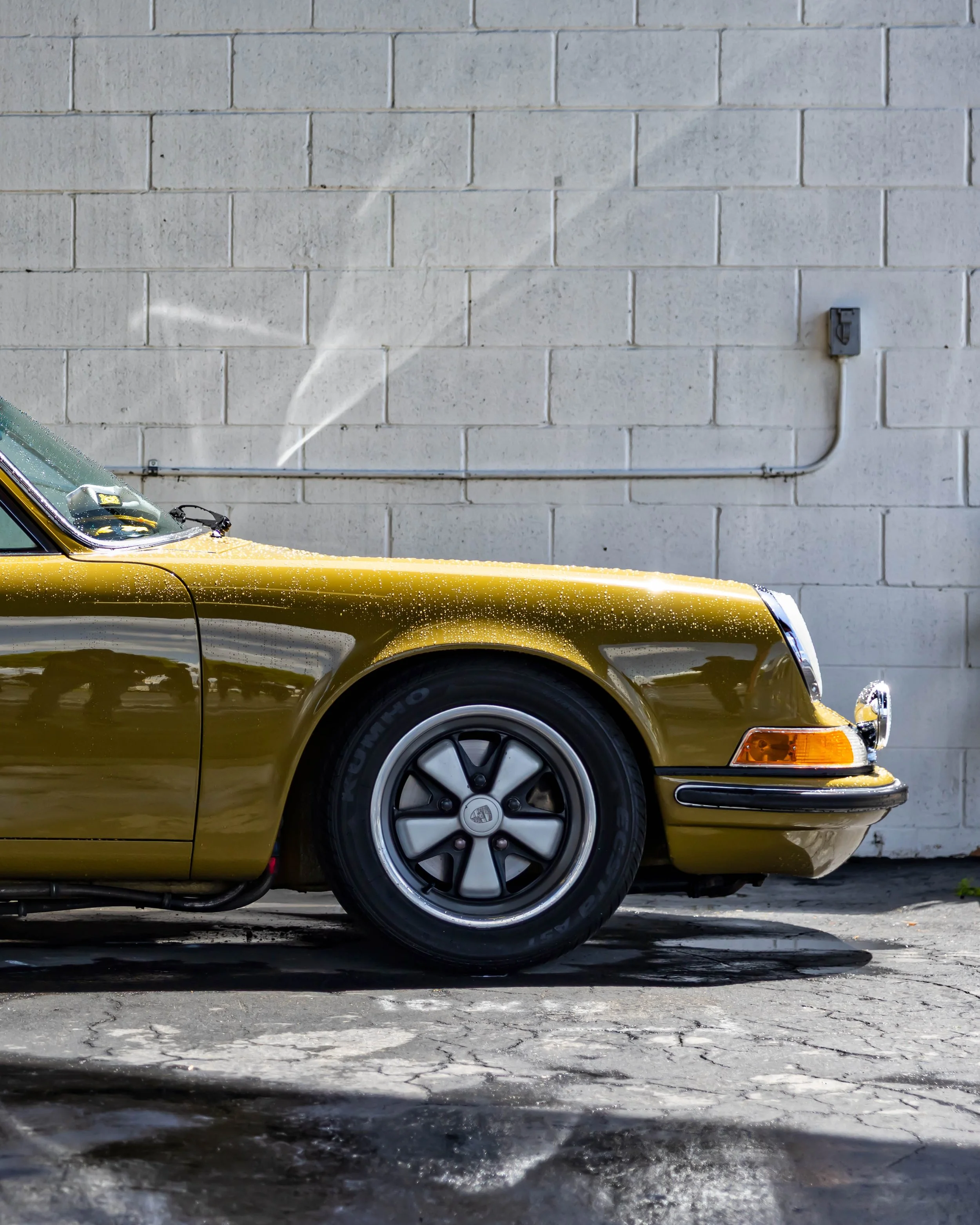 Air | Water 2025 — Orange County Fairgrounds — A low-angle, side profile view of the front quarter of a vintage Porsche 911 in a metallic olive green color. The car is covered in water droplets, suggesting it is being washed, and is parked on wet, cr