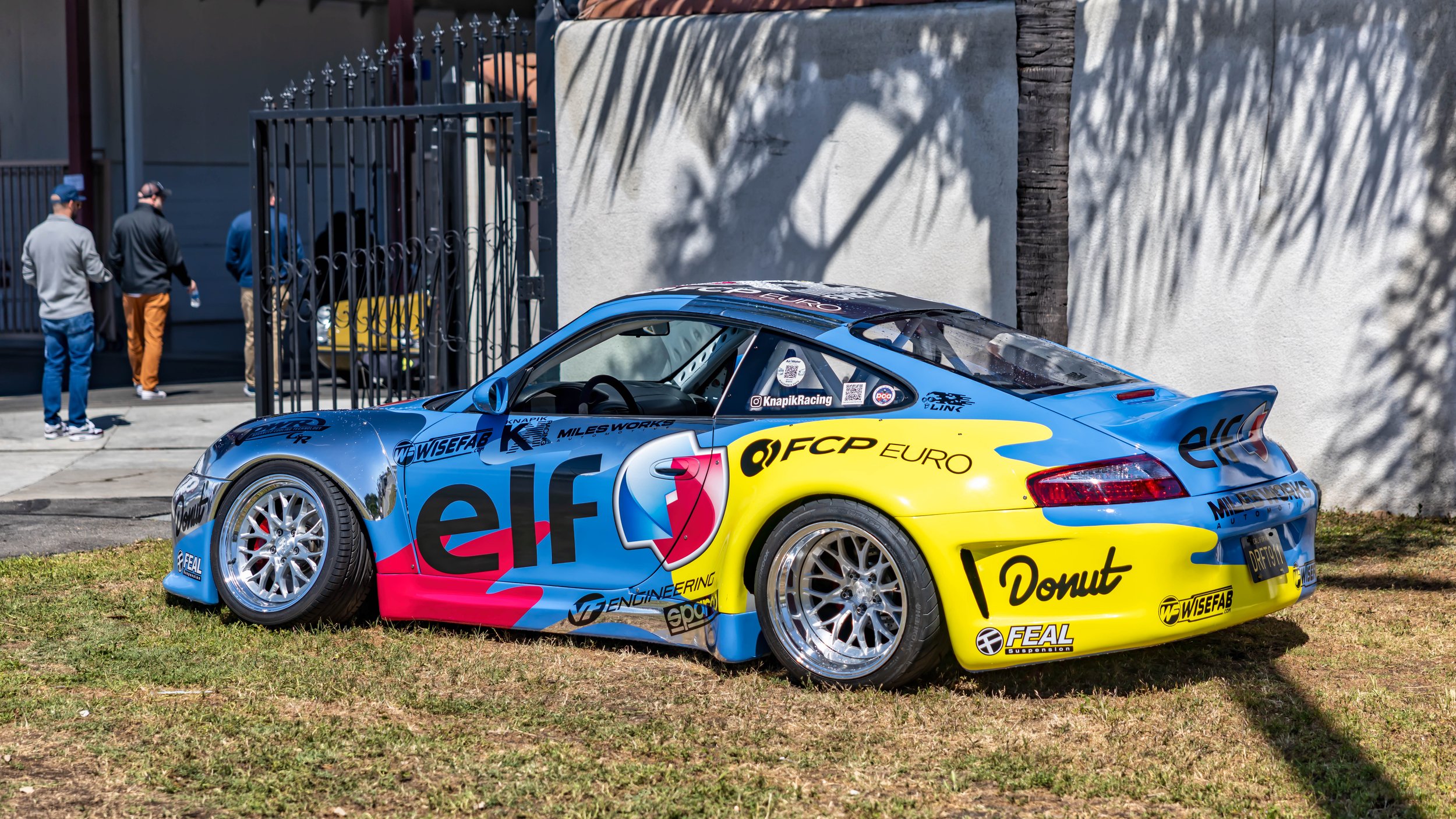 Air | Water 2025 — Orange County Fairgrounds — A heavily modified Porsche 911 (996) race car with a colorful blue, yellow, and pink livery featuring prominent 'elf', 'FCP Euro', and 'Donut' sponsorship decals. The car is parked on a grassy area and f