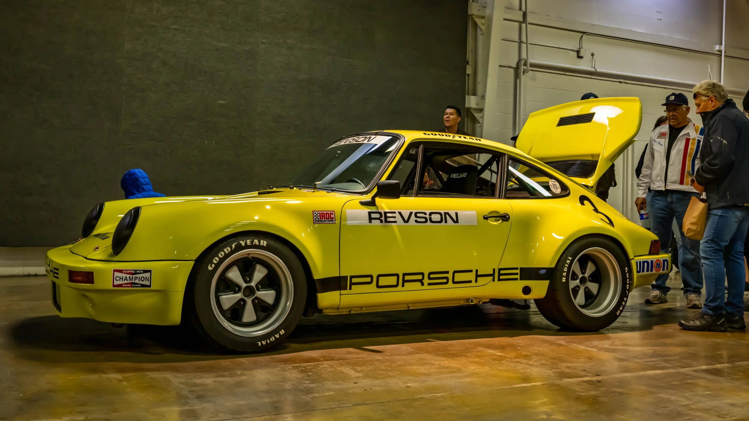Air | Water 2025 — Orange County Fairgrounds — A bright yellow 1974 Porsche 911 IROC RSR race car is parked indoors on a concrete floor, viewed from the front-left side. The car features a prominent 'REVSON' and 'PORSCHE' livery, the number '3' on th