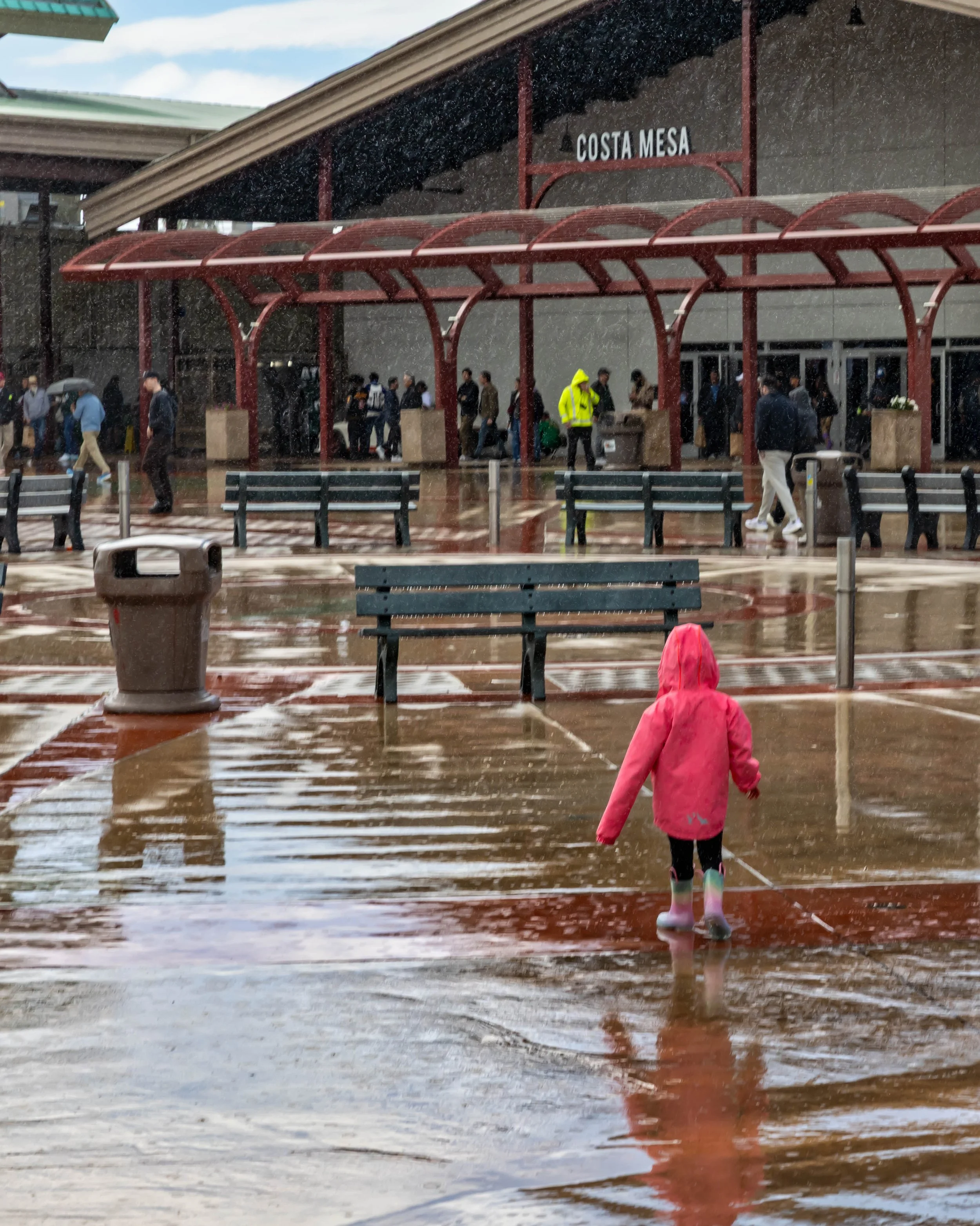 Air | Water 2025 — Orange County Fairgrounds — A young child in a bright pink hooded raincoat and colorful rain boots walks through a large puddle in a flooded plaza during a rain shower. In the background, other people are gathered under the covered