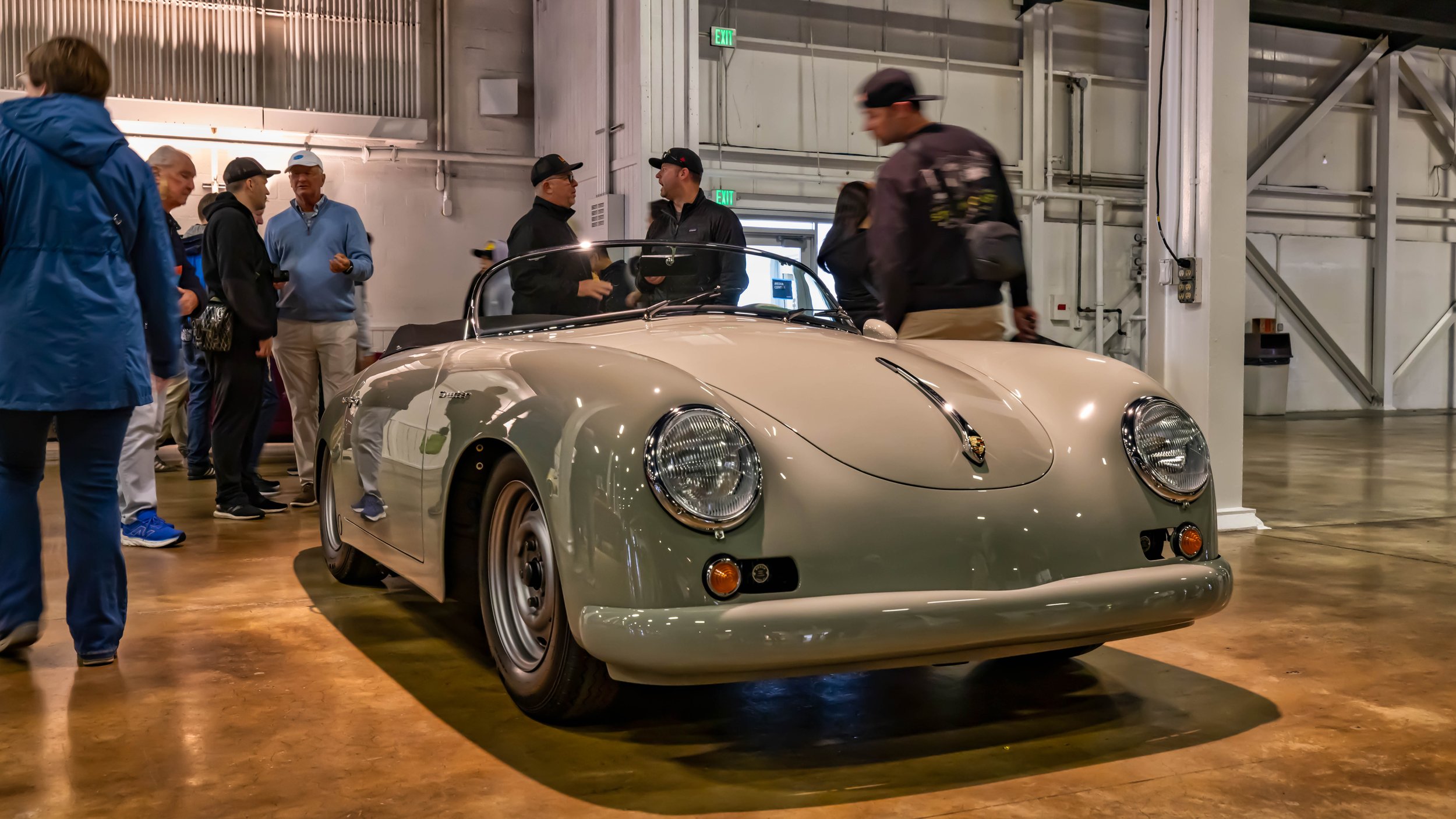 Air | Water 2025 — Orange County Fairgrounds — A classic Porsche 356 Speedster in a light stone gray color is parked indoors on a polished concrete floor. Spectators are gathered around the vehicle in a large, warehouse-like event space. — low angle,