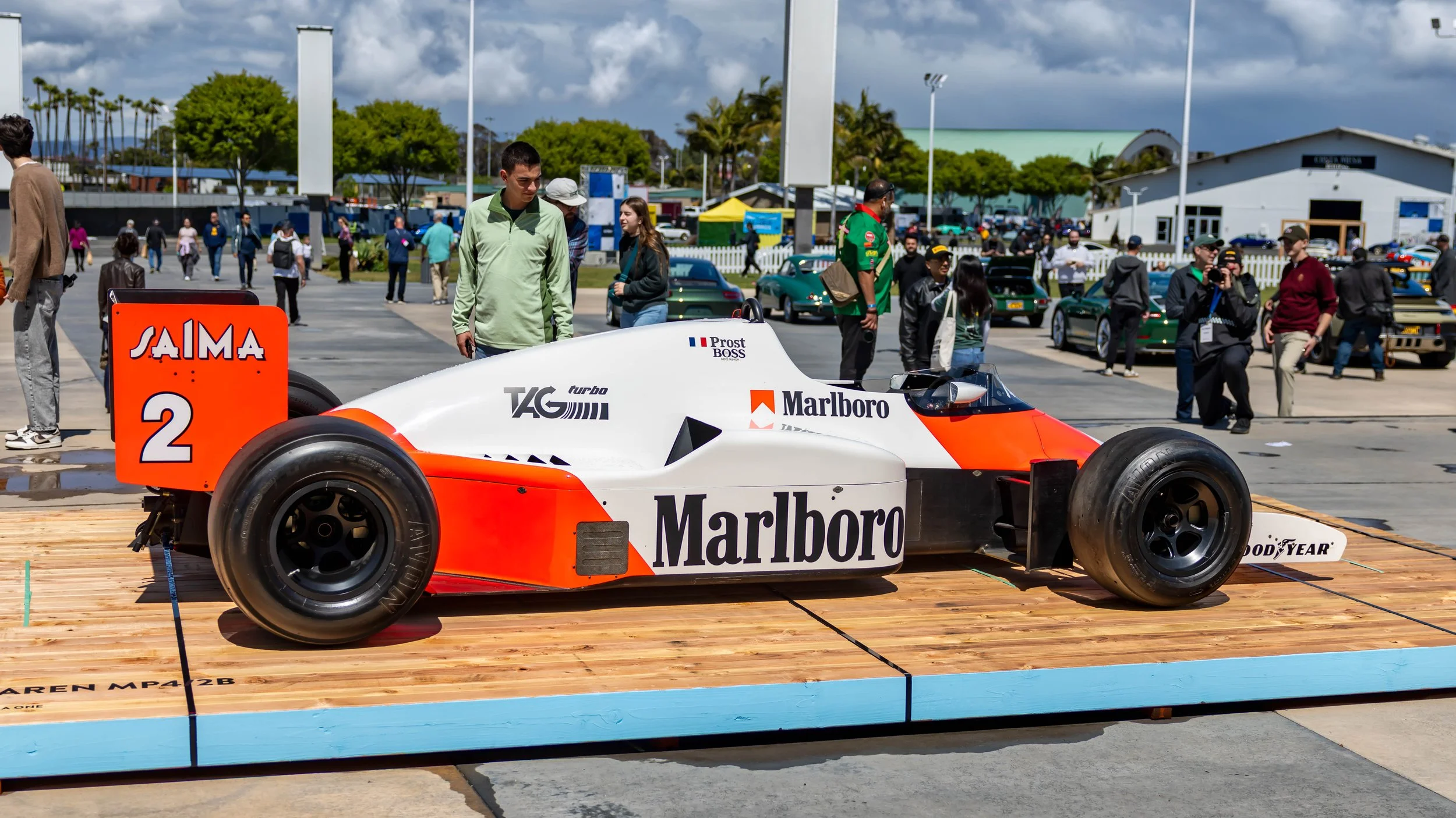 Air | Water 2025 — Orange County Fairgrounds — A 1985 McLaren MP4/2B Formula 1 car, featuring its iconic white and fluorescent red Marlboro livery, is displayed on a wooden platform at an outdoor car event. Spectators are gathered around the historic