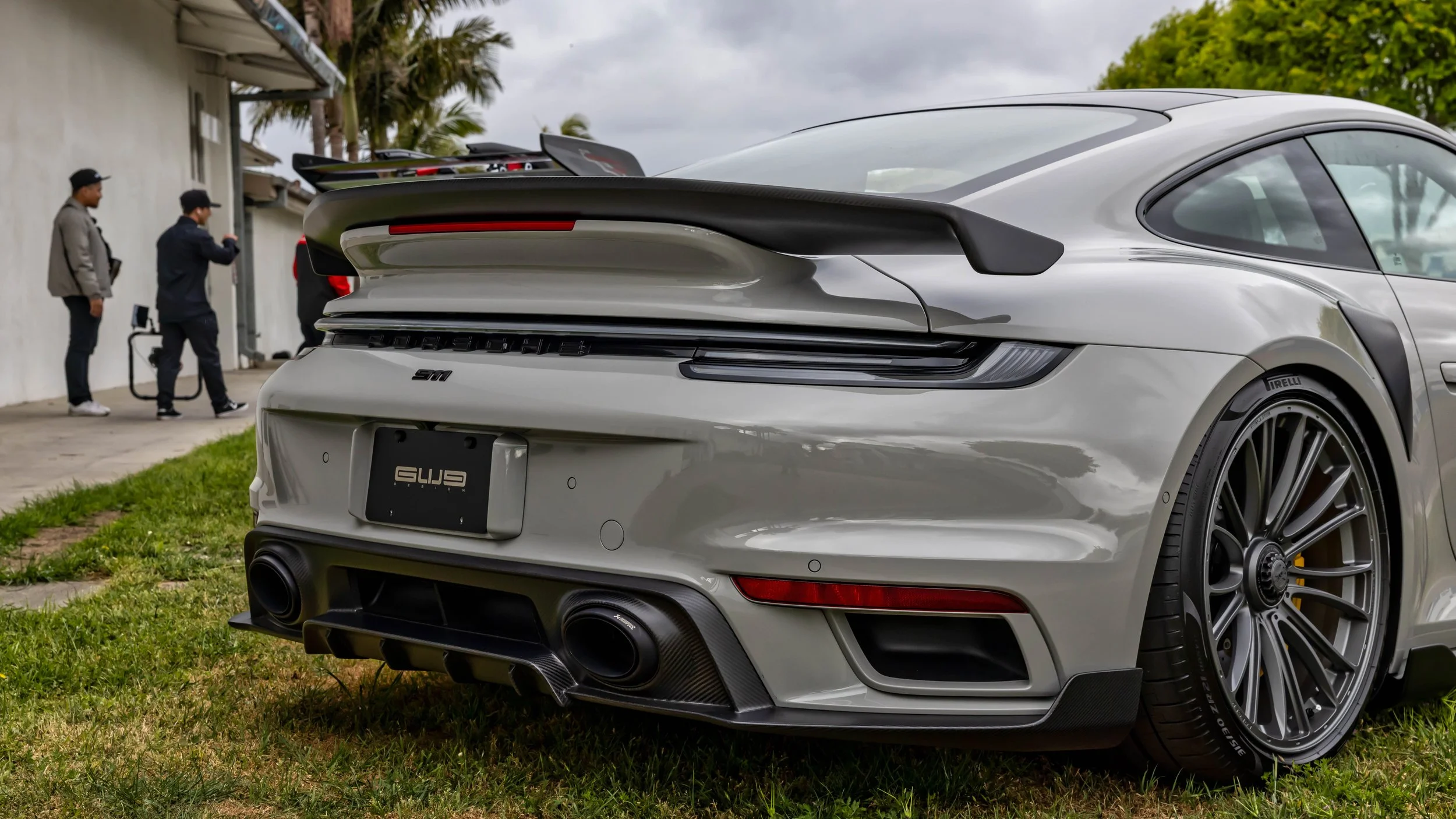 Air | Water 2025 — Gunther Werks — Orange County Fairgrounds — A low-angle, rear three-quarter view of a Chalk Grey Porsche 911 Turbo S (992) parked on a grassy area next to a building. The car features GW9 992 carbon-fiber aero kit with this shot ca