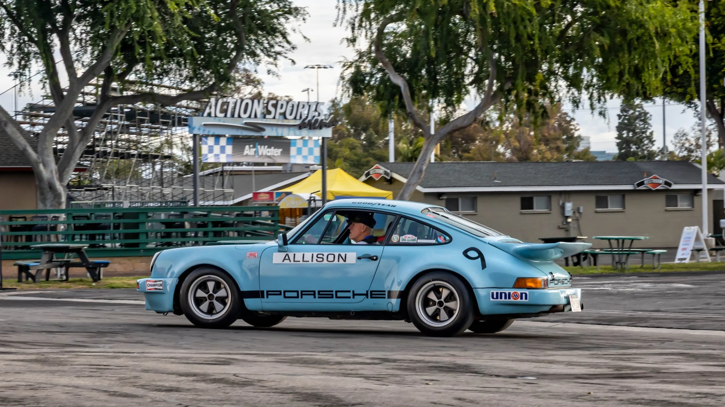 Air | Water 2025 — Orange County Fairgrounds — A light blue, race-prepared classic a a 1974 Porsche 911 RSR IROC tribute car, adorned with "PORSCHE" side livery and the number 9, is being driven by a man in a paved lot. The car features a prominent "