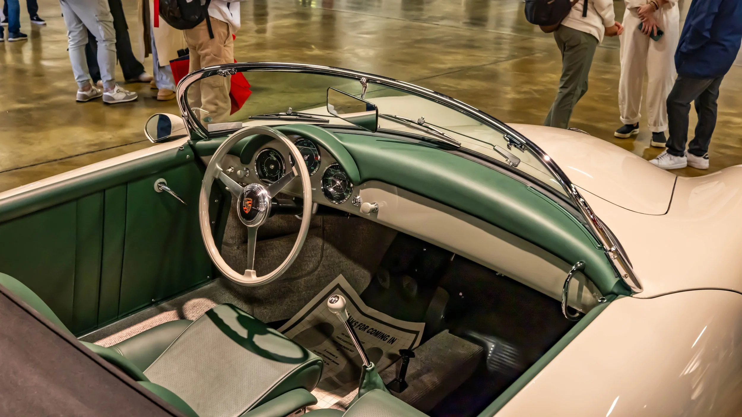 Air | Water 2025 — Orange County Fairgrounds — A high-angle view into the driver's side cockpit of a classic Porsche 356 Speedster, showcasing its dark green leather interior against a cream-colored exterior. The minimalist dashboard features three r