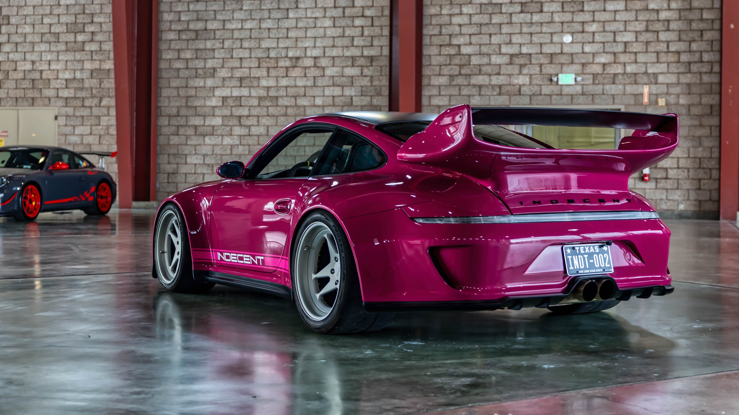 Air | Water 2025 — Orange County Fairgrounds — A low-angle, three-quarter rear view of a heavily modified, widebody Porsche 997 in a vibrant magenta color. The car features a large rear wing, deep-dish silver five-spoke wheels, and 'INDECENT' livery 