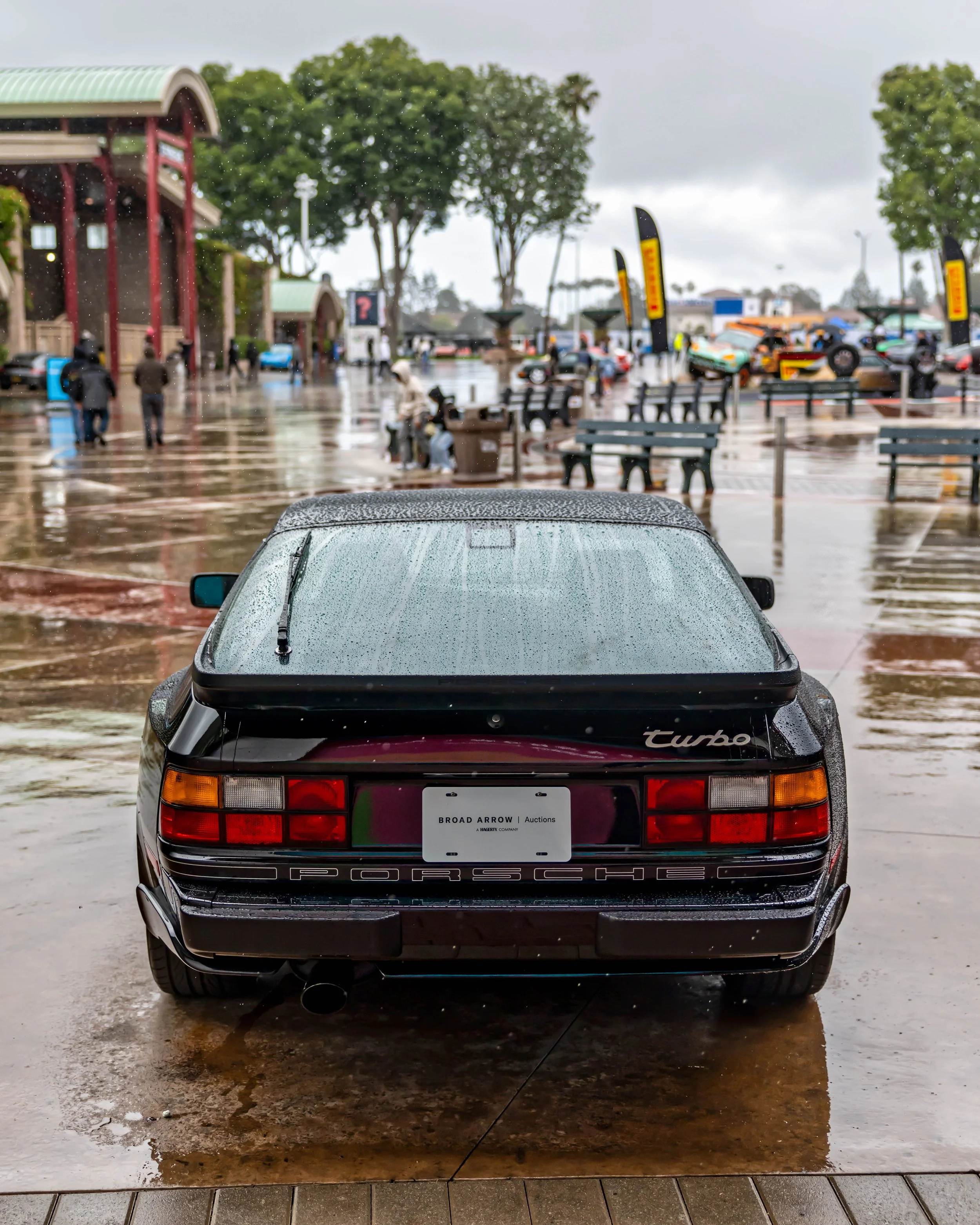 Air | Water 2025 — Orange County Fairgrounds — A rear view of a black Porsche 944 Turbo parked on a wet, paved plaza during a rainy day. The car's body and rear window are covered in raindrops, with reflections visible on the glistening ground. In th
