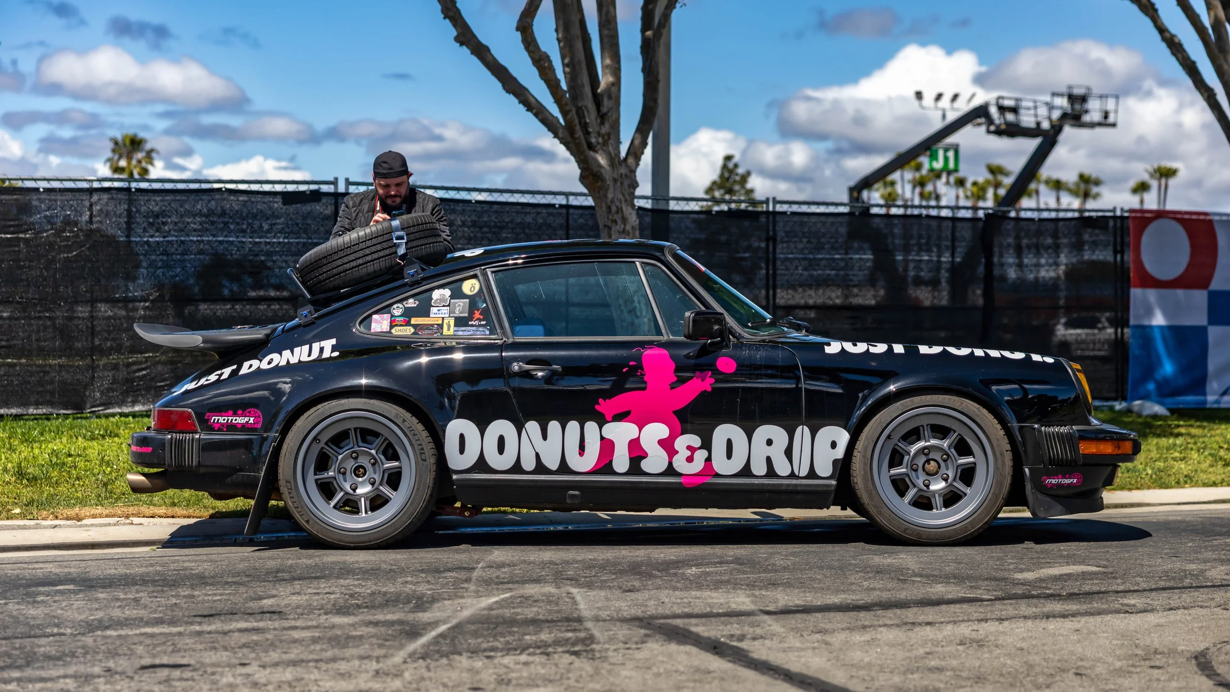 Air | Water 2025 — Orange County Fairgrounds — A man in a black cap secures a spare tire to a roof rack on a black Porsche 911 G-body. The car is heavily customized with a large white and pink 'DONUTS & DRIP' livery, a 'whale tail' spoiler, and grey 