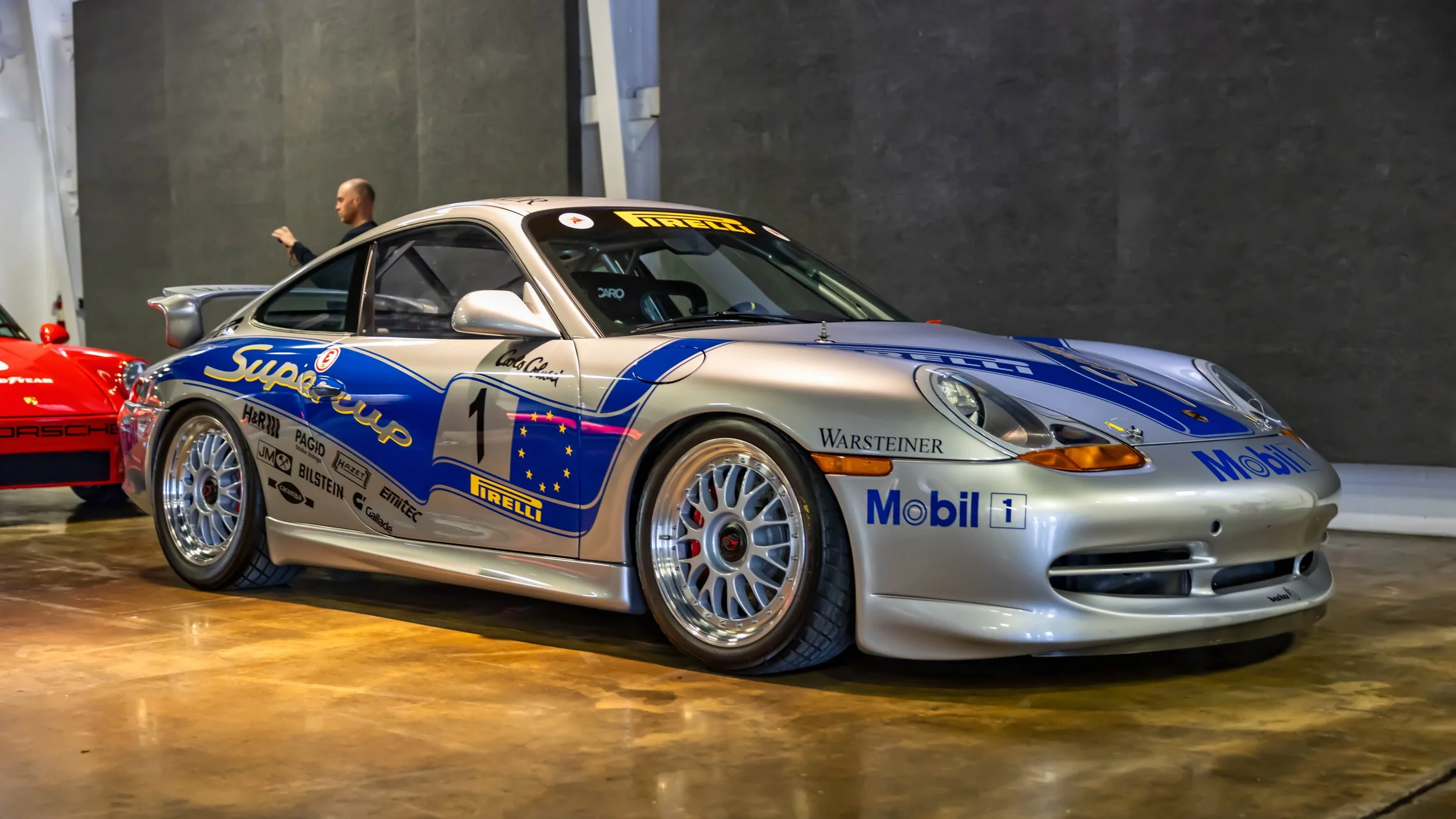 Air | Water 2025 — Orange County Fairgrounds — A silver Porsche 996 GT3 Cup car with a prominent blue and yellow Mobil 1 Supercup livery is parked indoors on a polished concrete floor. The car, bearing the number 1, is covered in period-correct spons