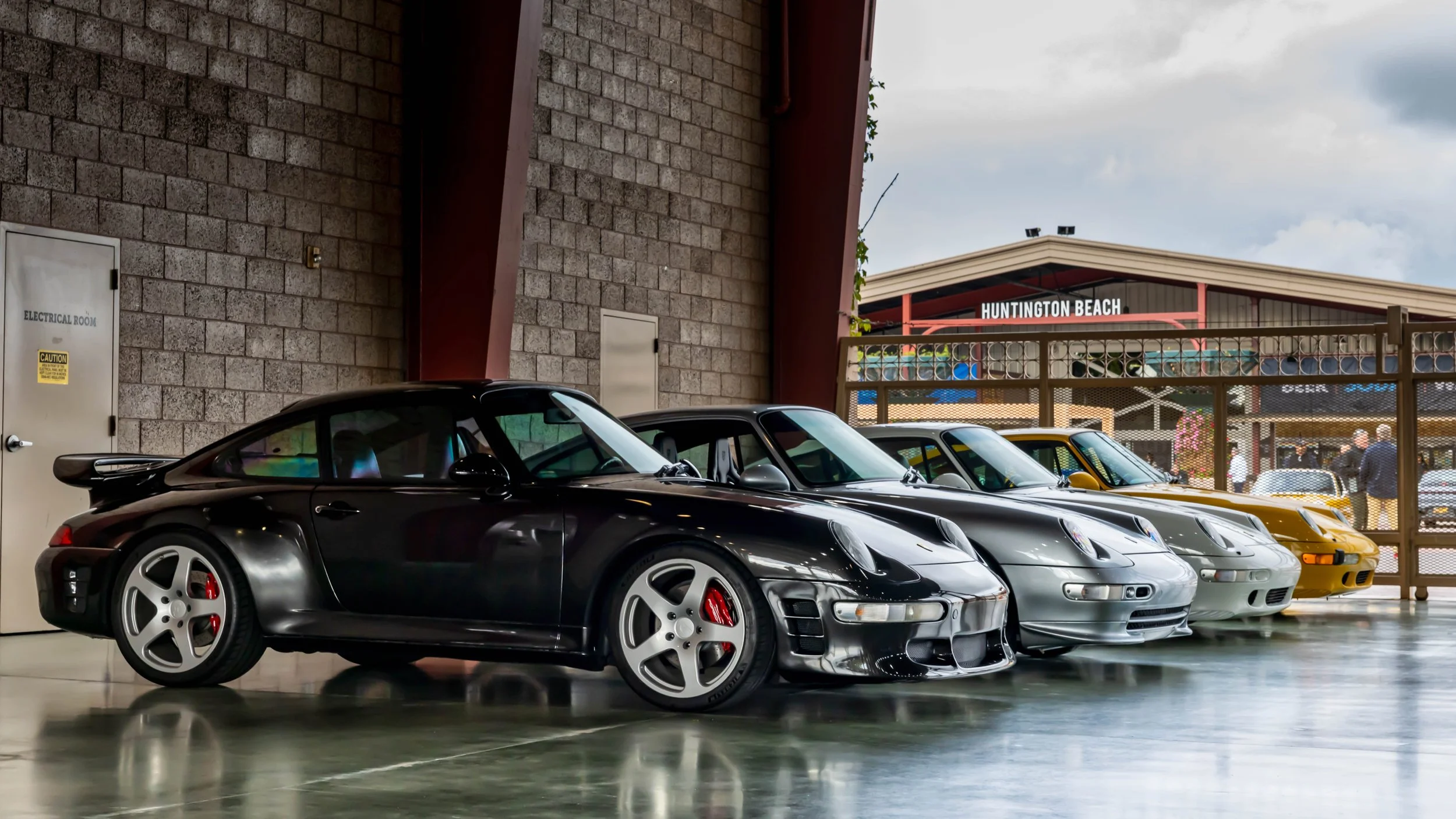 Air | Water 2025 — Orange County Fairgrounds — A lineup of several RUF 911s, led by a black RUF CRT2, is parked indoors on a reflective concrete floor. The lead car features distinctive 5-spoke wheels and red brake calipers. In the background, other 