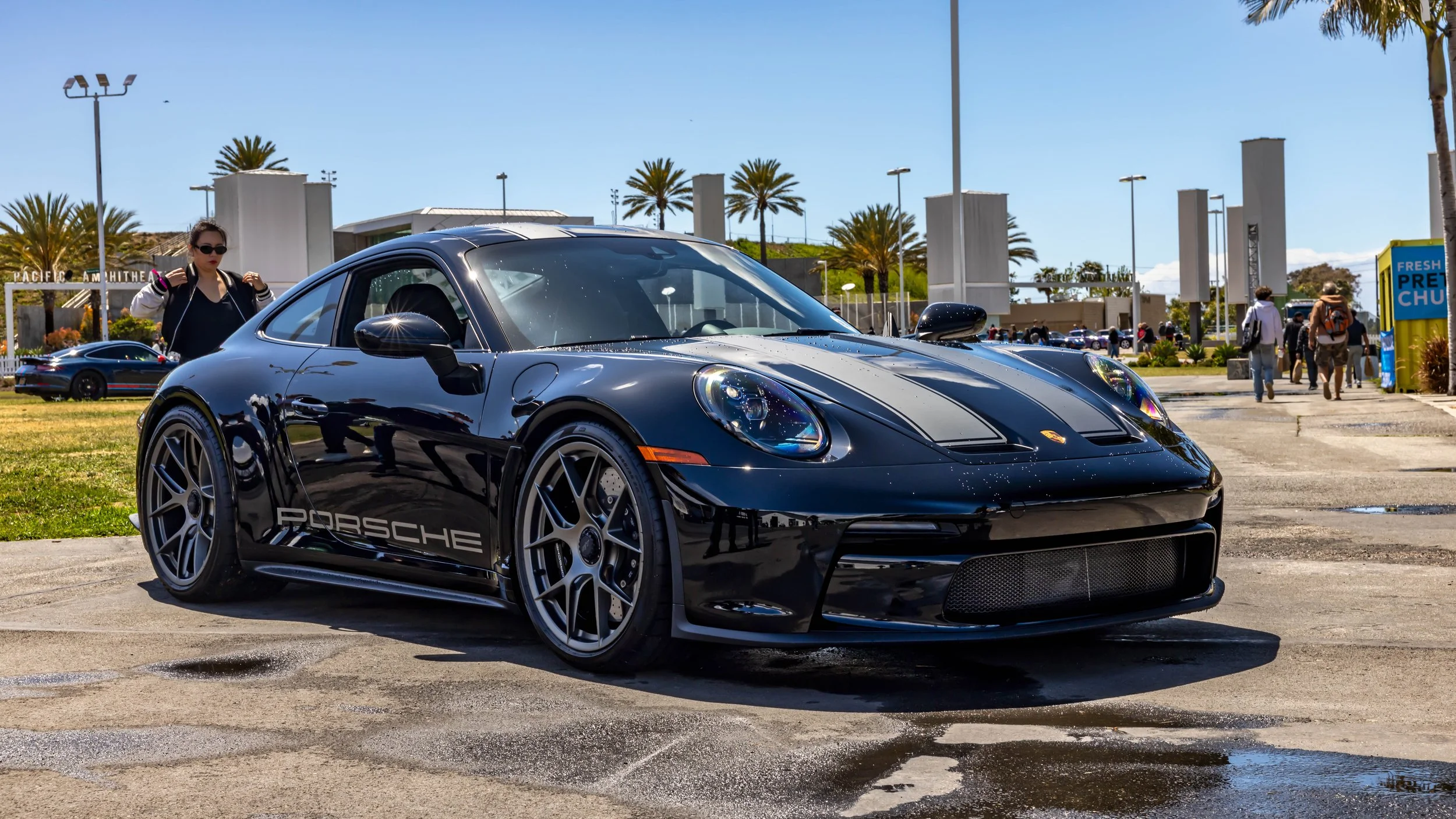 Air | Water 2025 — Orange County Fairgrounds — A black Porsche 911 S/T (992) with grey racing stripes and side decals is parked on a wet patch of pavement at an outdoor event. The car is shown from a low front-three-quarters angle, highlighting its f