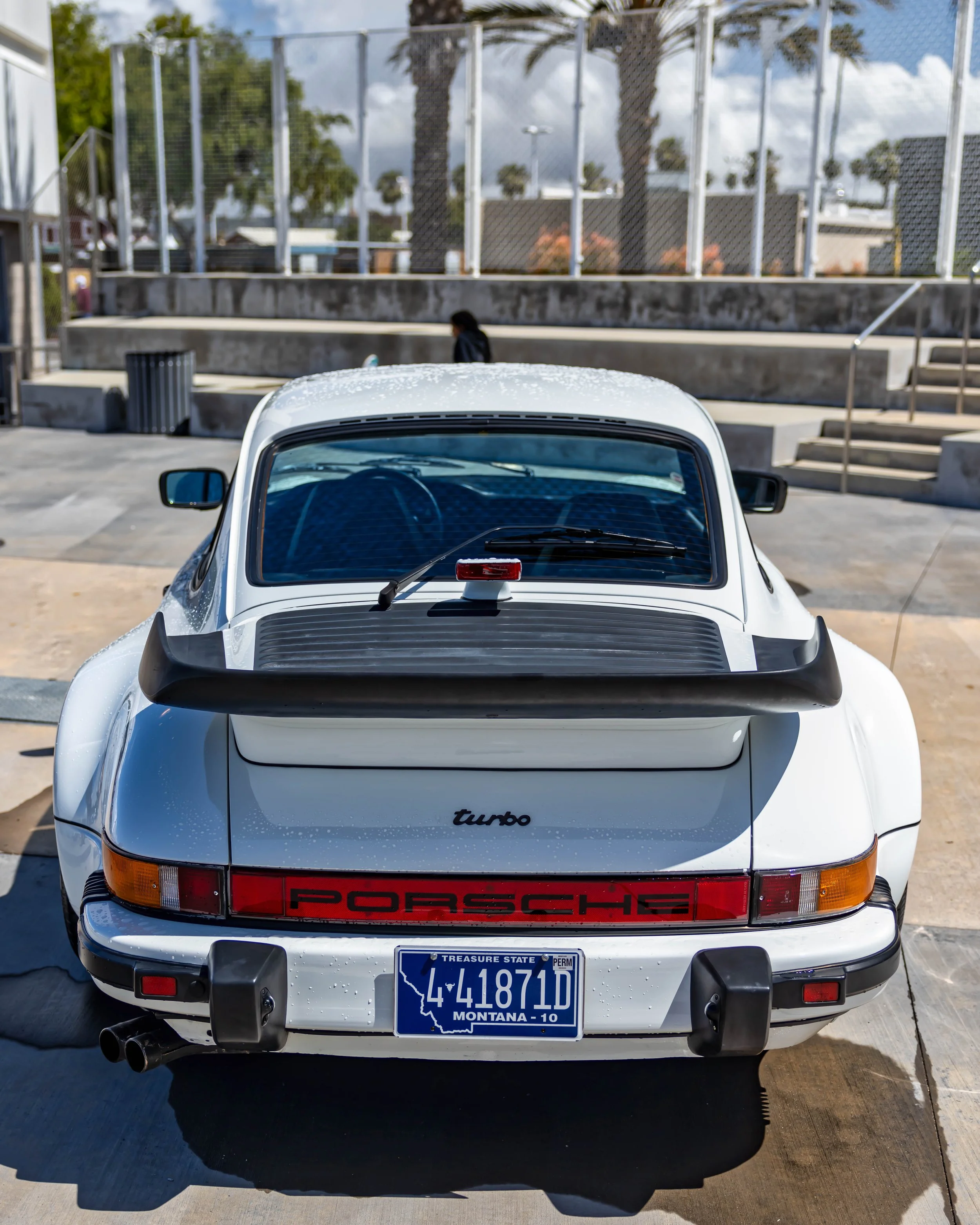 Air | Water 2025 — Orange County Fairgrounds — A rear view of a white G-body Porsche 911 Turbo (930), distinguished by its wide rear fenders and large 'whale tail' spoiler. The car is covered in water droplets and is parked on a concrete surface, wit