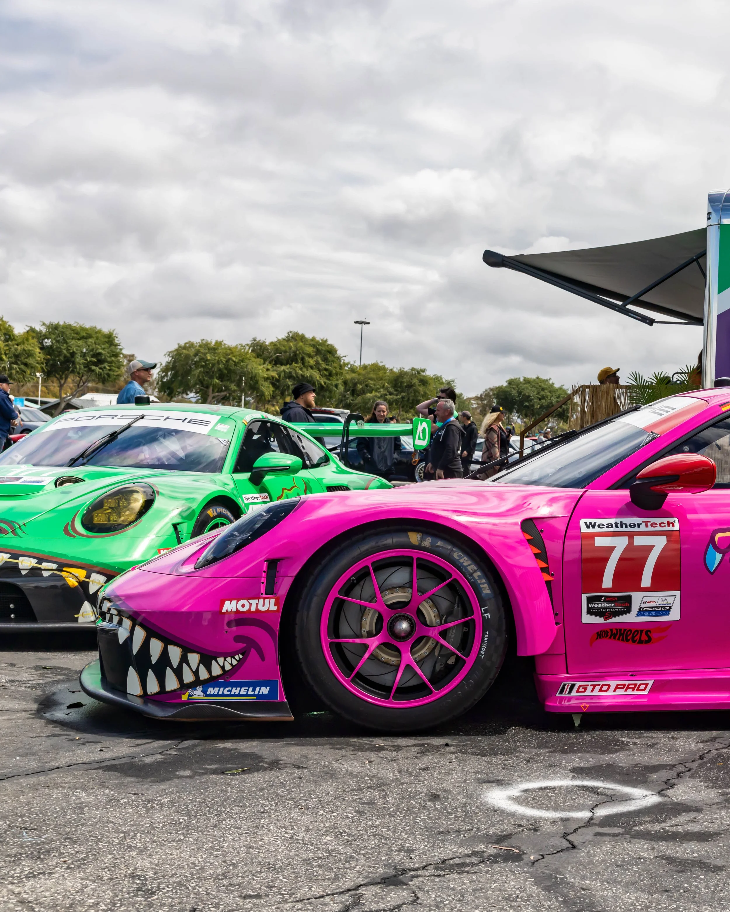 Air | Water 2025 — Orange County Fairgrounds — A low-angle, close-up shot of a vibrant pink #77 Porsche 911 GT3 R race car, featuring a prominent shark-mouth livery on its front bumper. The car is parked on asphalt in a paddock setting, with a simila