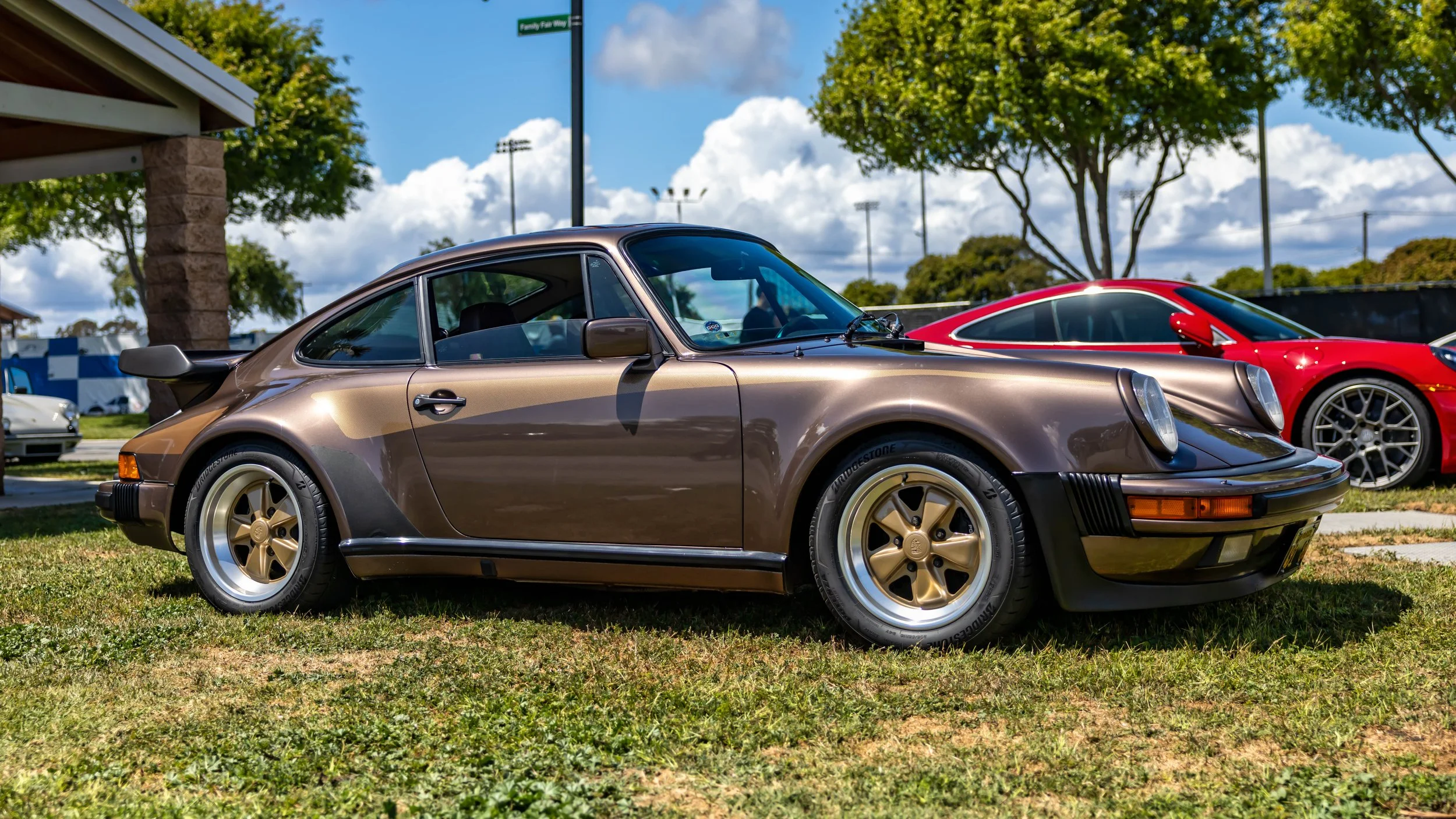 Air | Water 2025 — Orange County Fairgrounds — A low-angle, front-three-quarter view of a vintage Porsche 911 Turbo (930) in a metallic brown finish with gold side graphics. The car is parked on a grassy lawn under a bright, partly cloudy sky. It fea