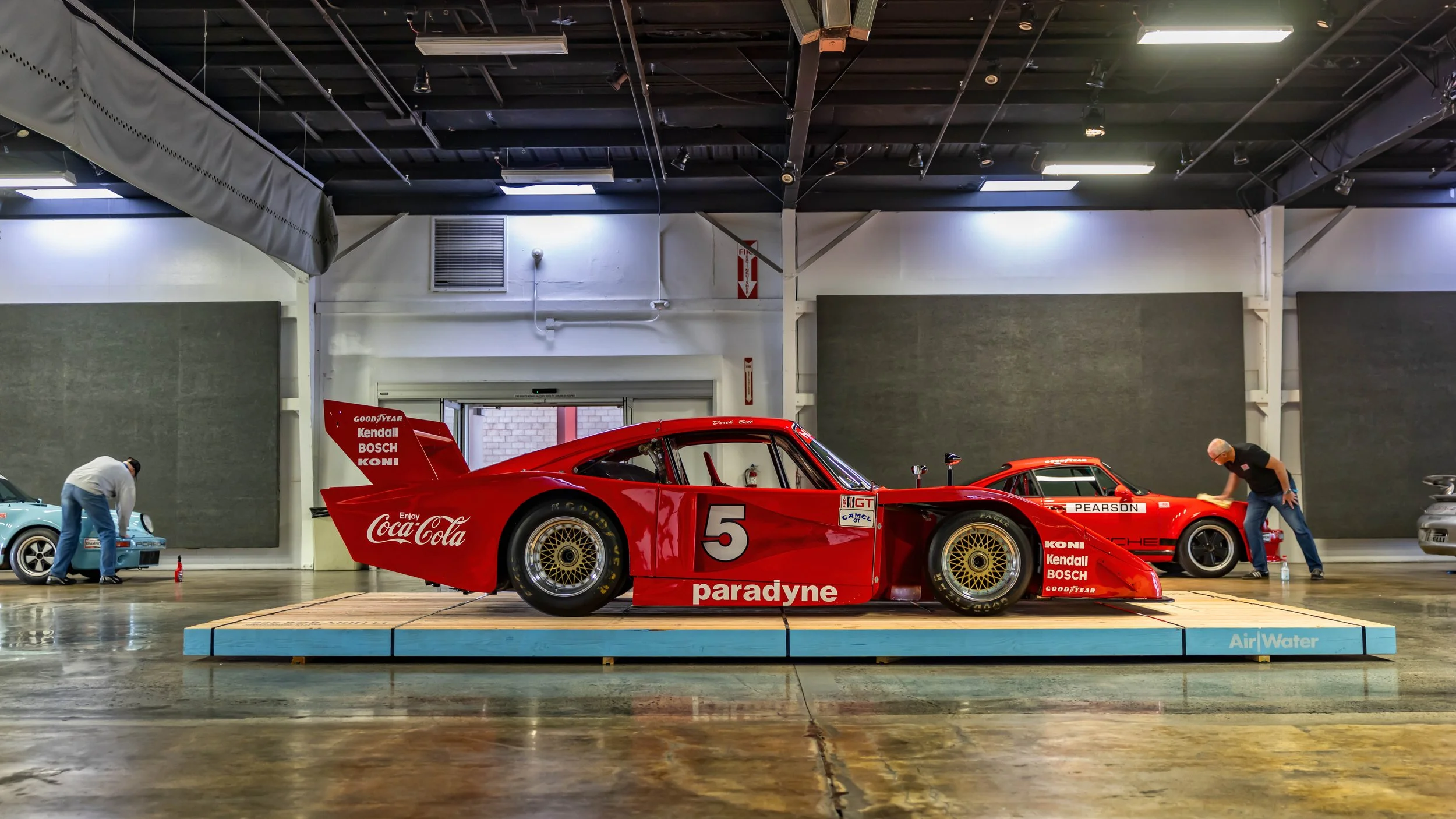 Air | Water 2025 — Orange County Fairgrounds — A low-angle, profile view of the iconic red #5 Bob Akin Racing Porsche 935 K3, featuring its distinctive Coca-Cola and Paradyne livery. The race car is elevated on a light blue and wood display platform 