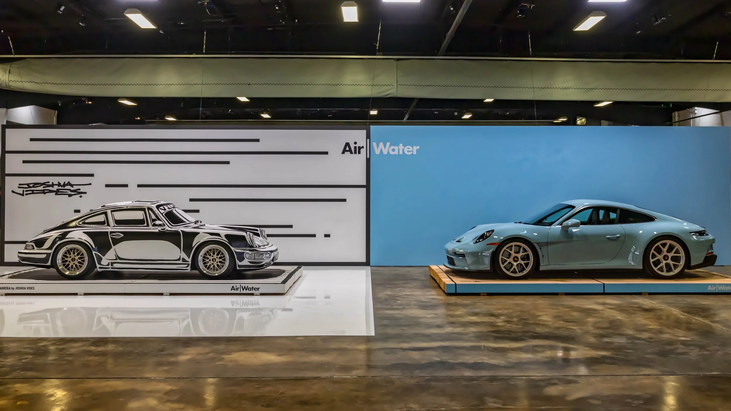 Air | Water 2025 — Orange County Fairgrounds — A comparative display at an 'Air | Water' event, featuring two Porsche 911s side-by-side. On the left, a classic air-cooled 911 Carrera is presented as a black-and-white, two-dimensional art piece by Jos